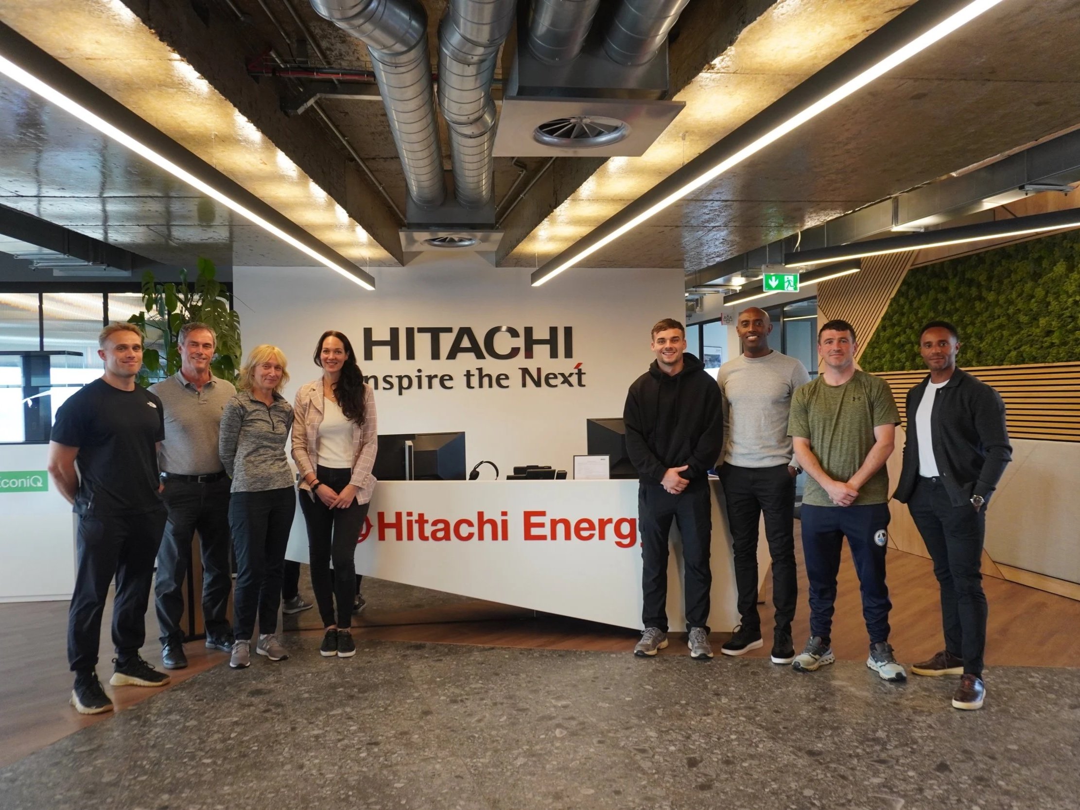 Group of eight people standing in front of a reception desk at Hitachi, with the company's logo and slogan in the background.
