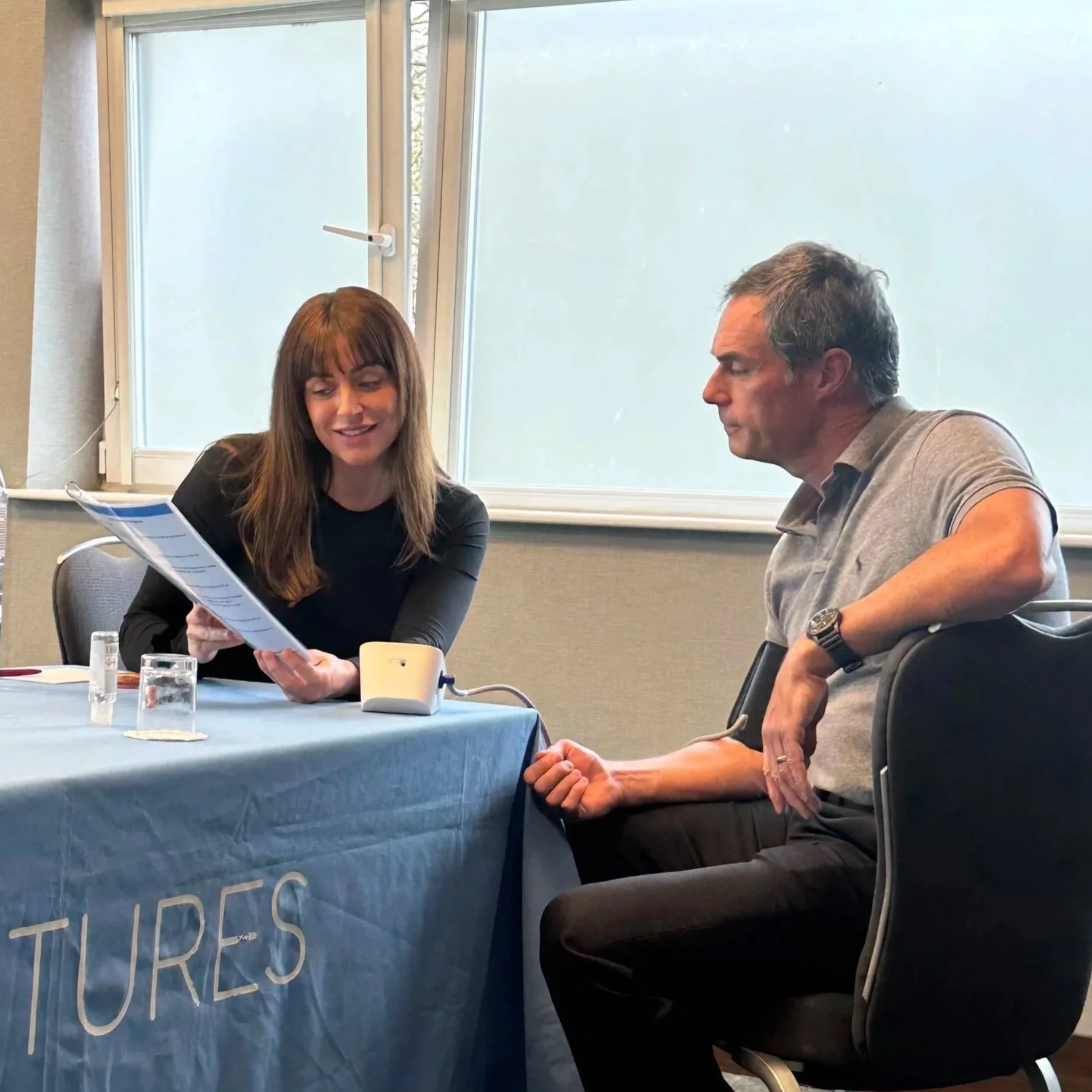 A woman and an older man sitting at a table engaged in conversation during a meeting. The woman is holding a document and smiling, while the man is listening attentively with his arm resting on the table. The table has a blue tablecloth with partially visible text.