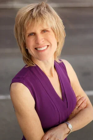A smiling woman with short blonde hair, wearing a purple sleeveless top and a watch, standing with her arms crossed in front of a neutral background.