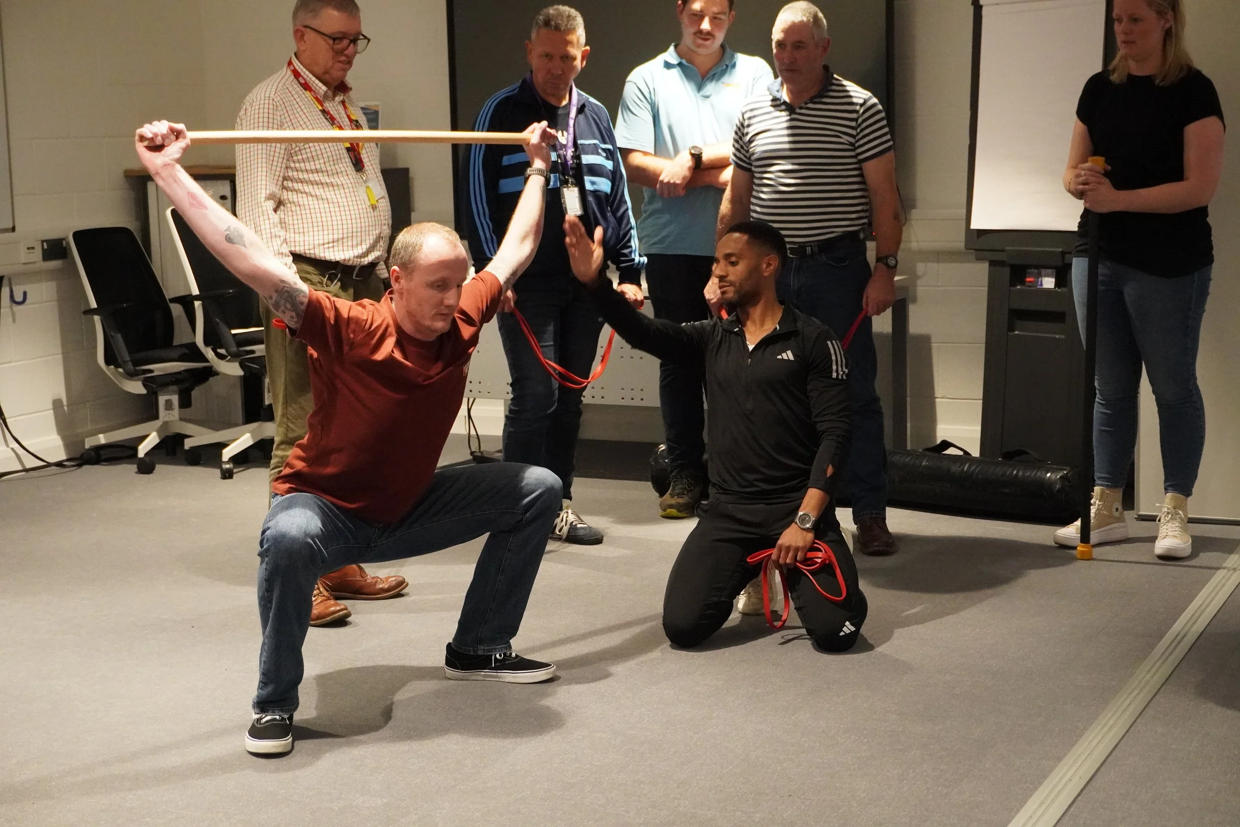 Group of six people in a room participating in a physical training exercise with resistance bands, with three men in front and three women in the background observing.