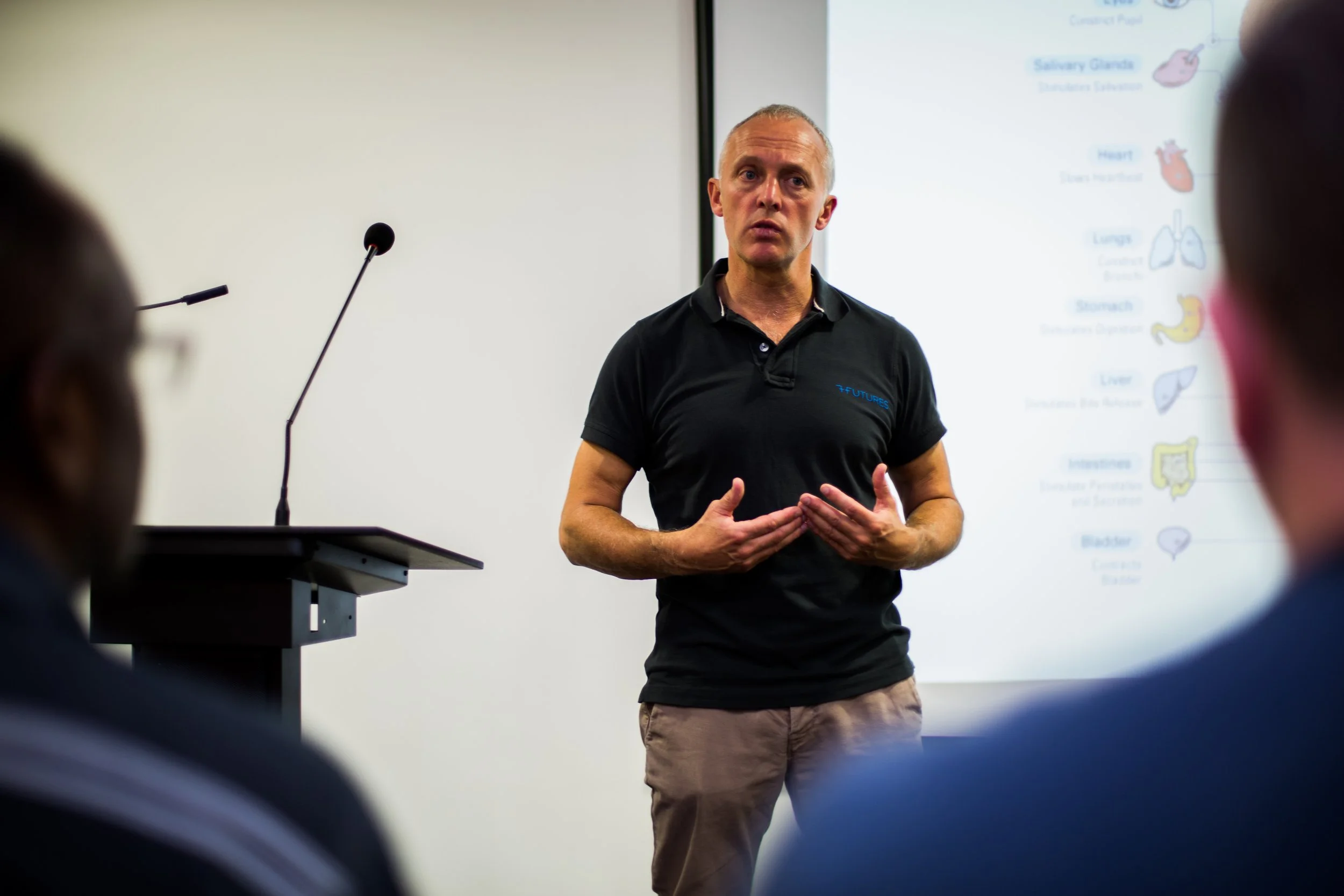 A man giving a presentation to an audience in a conference room, with a screen displaying a diagram of human organs behind him.