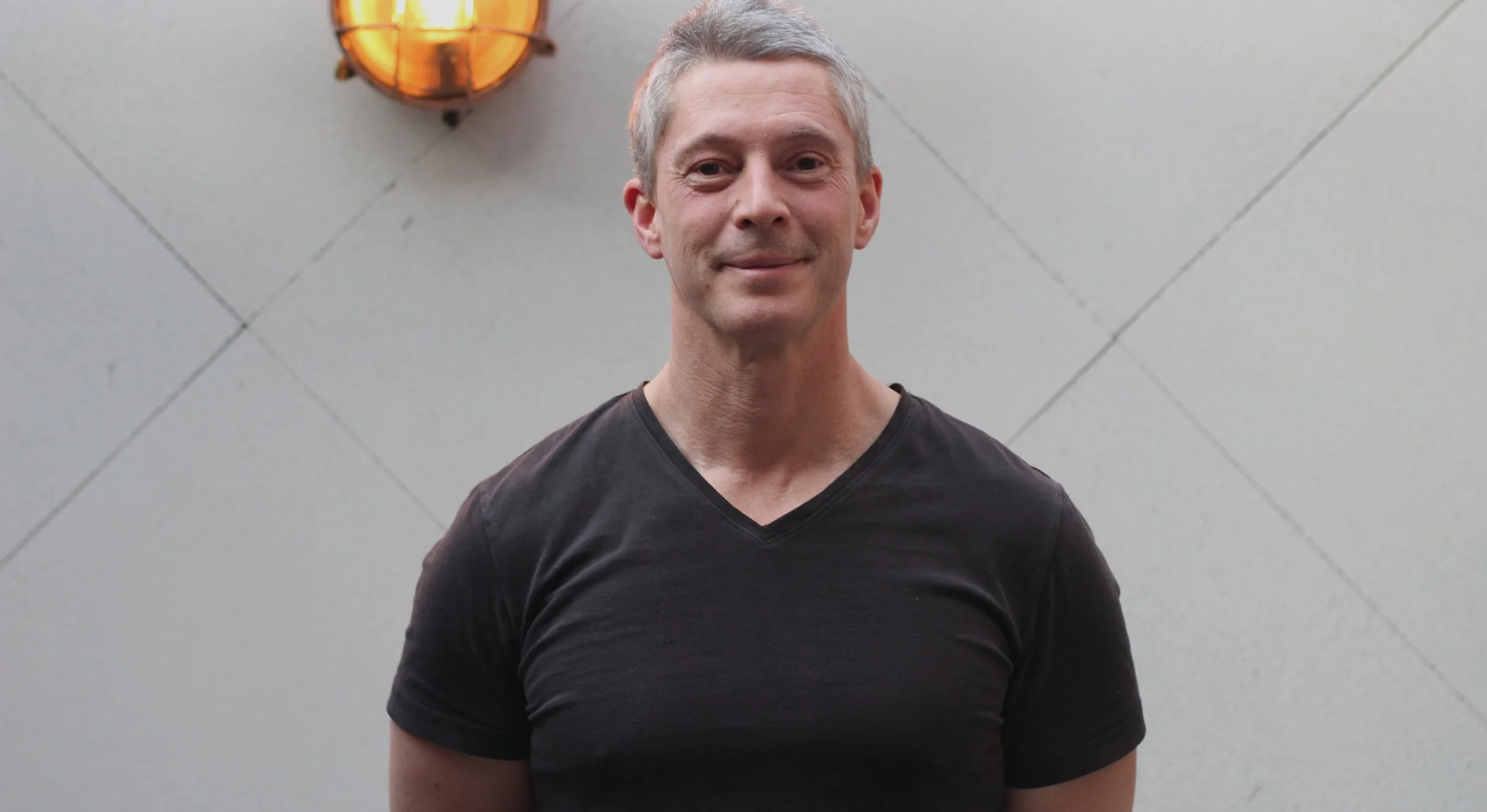A middle-aged man with short gray hair wearing a black V-neck shirt, standing against a plain tiled wall with an orange light fixture in the background.