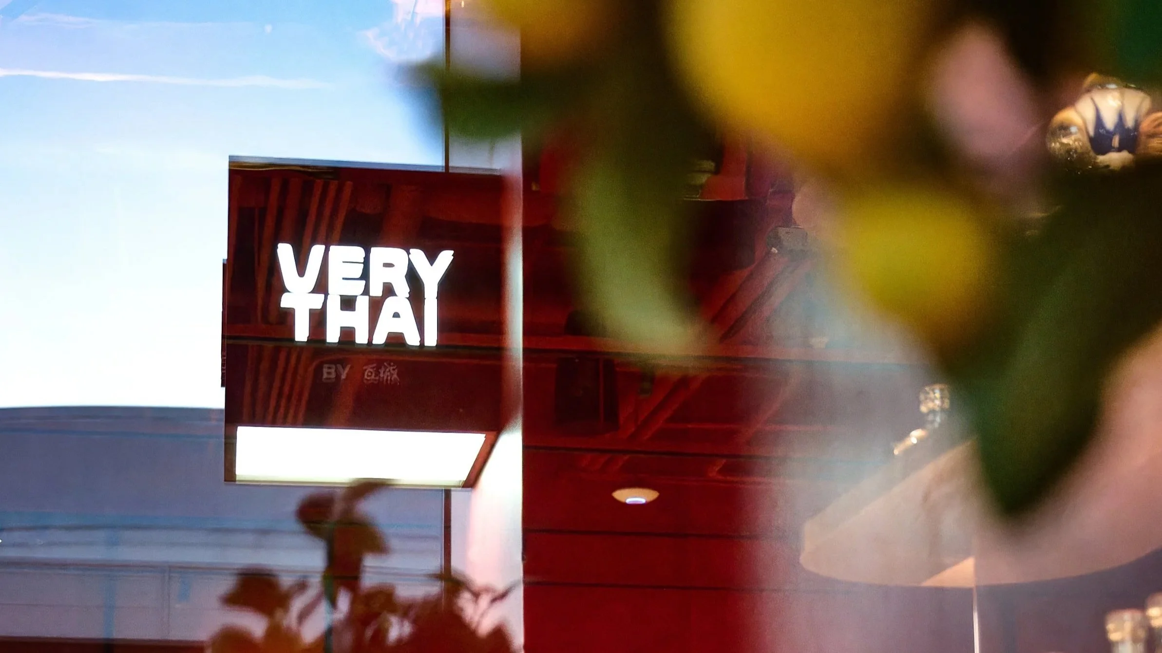 Reflective glass surface shows a brightly lit sign that reads 'VERY THAI' in white letters against a dark background, with visible cloud sky and part of an indoor ceiling with wooden beams and objects, partially obscured by colorful out-of-focus refl