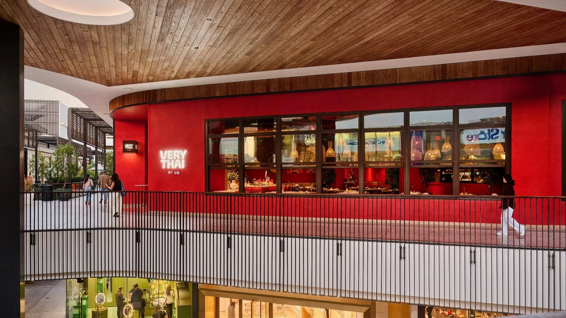 View of a modern shopping mall with a red restaurant named 'Very Thai' on the upper floor, featuring large windows, warm interior lighting, and a curved wooden ceiling. People are walking on the walkway, and a railing separates the walkway from the l
