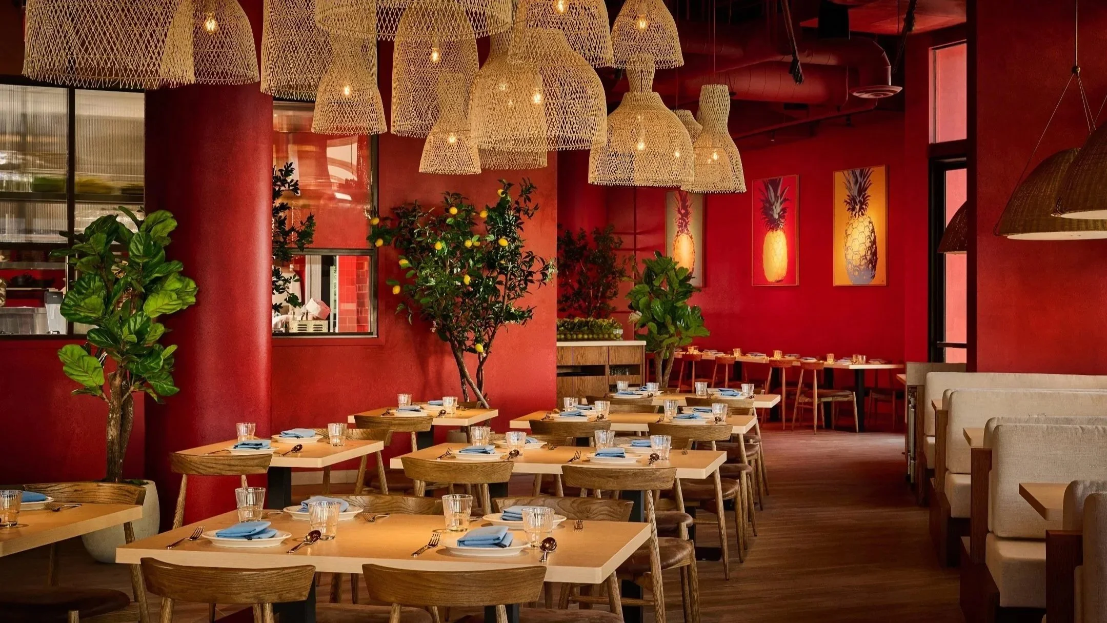 A stylish restaurant interior with red walls, wooden tables, and chairs. Hanging woven light fixtures, potted green plants, framed pineapple artwork, and a row of booths along the right side.