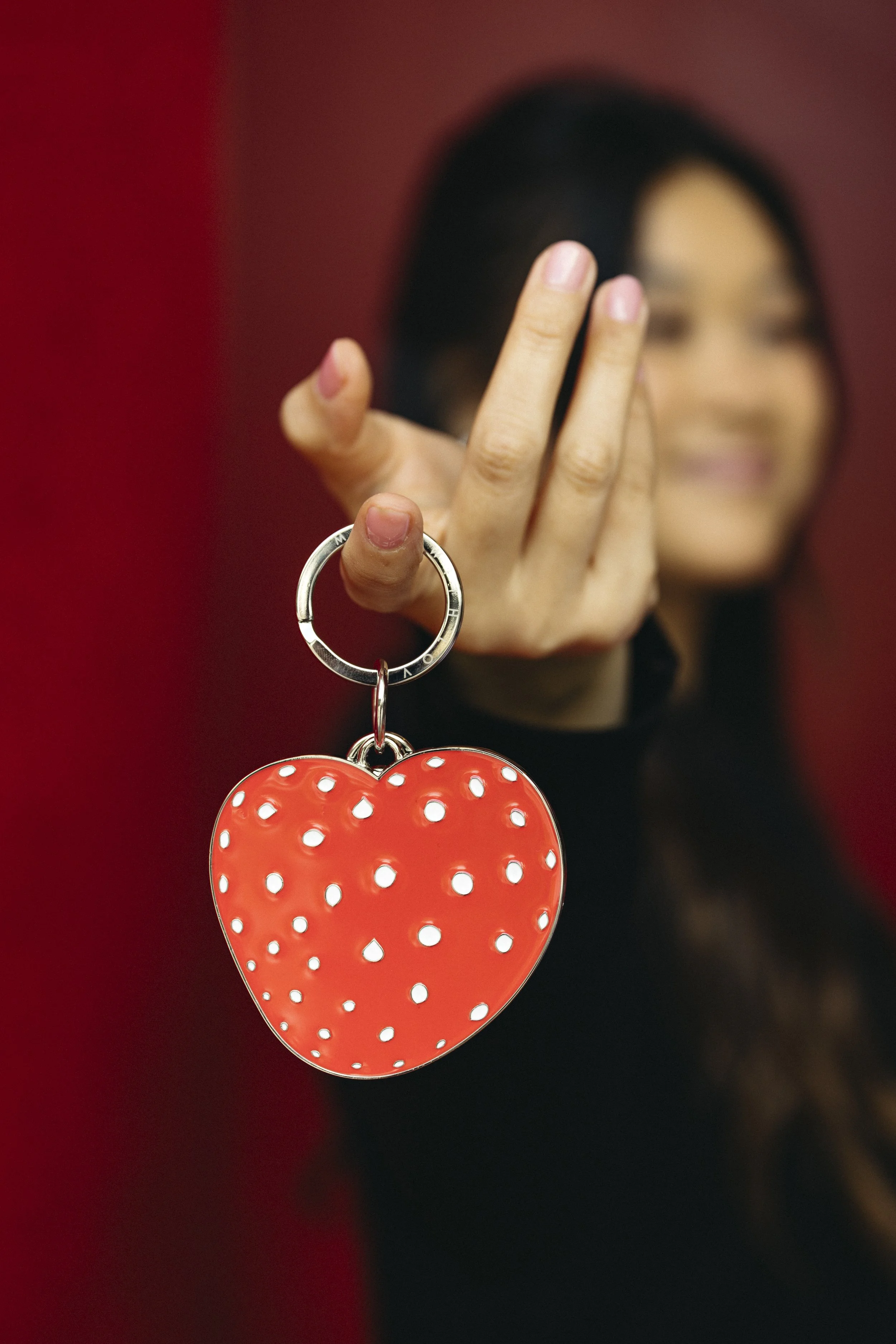 A woman with glasses and dark hair holding a red heart-shaped keychain with white polka dots in front of her face, against a red background.