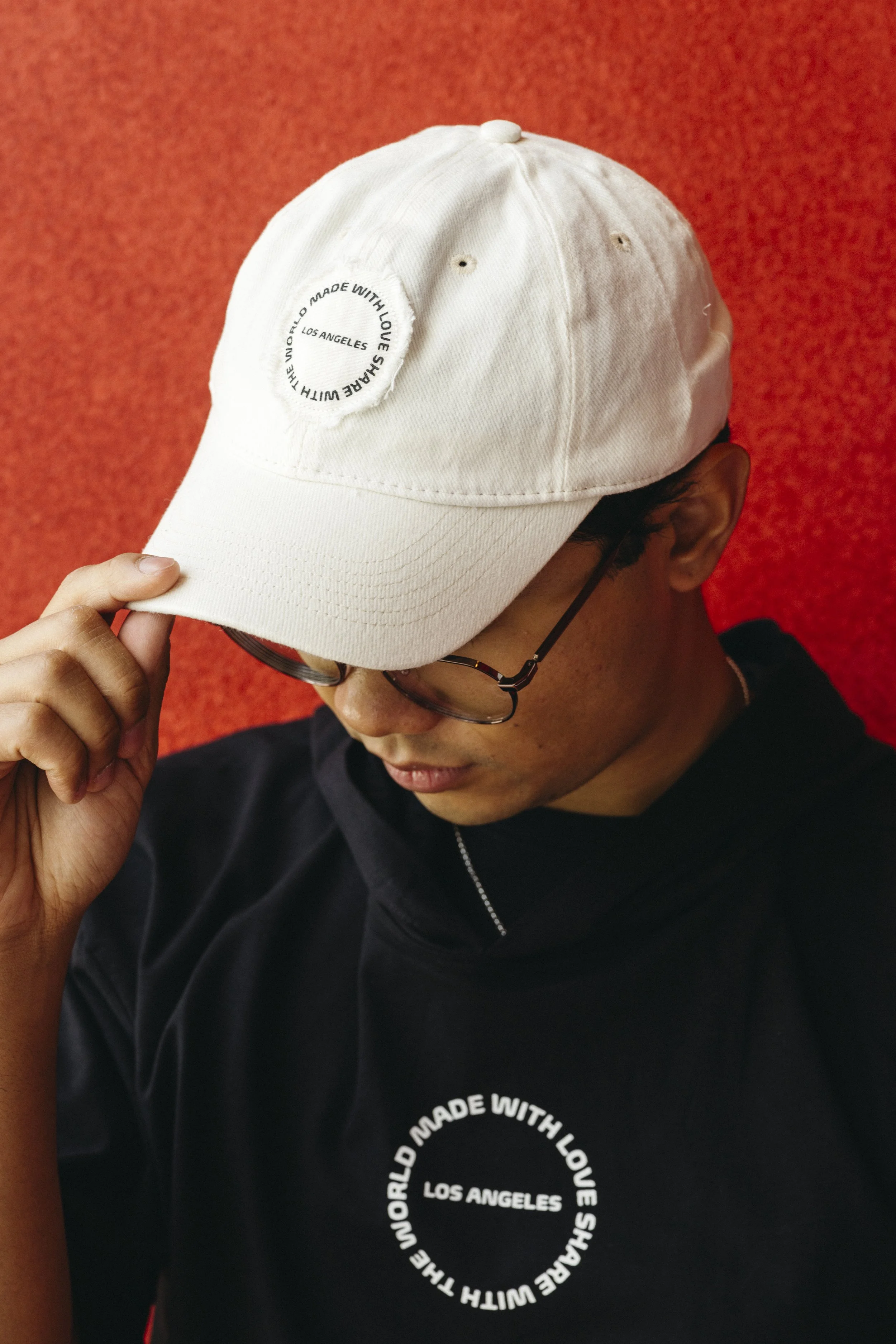 Person wearing glasses and a black hoodie with a white baseball cap covering their face, against a red background.