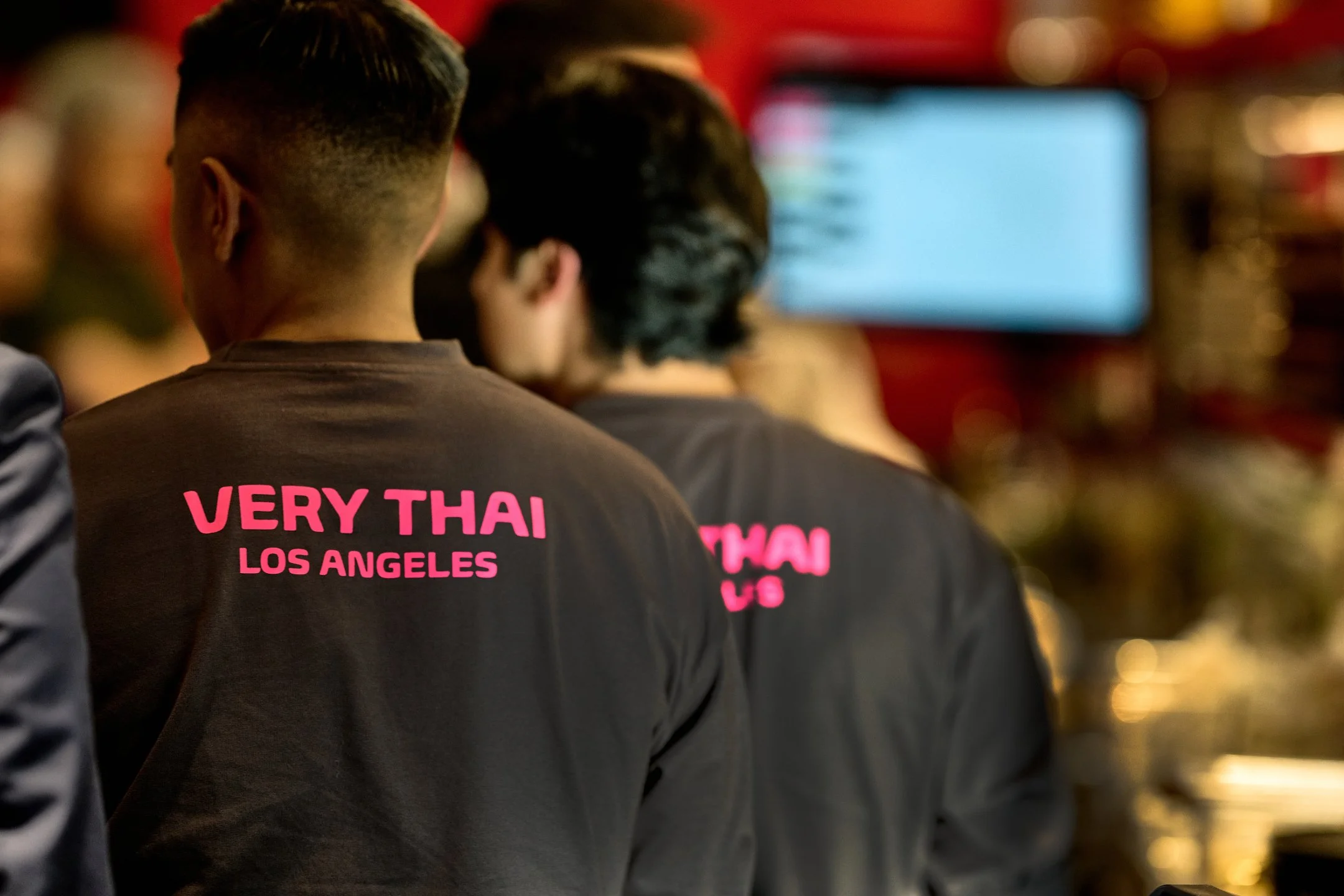 People wearing black t-shirts with pink text reading 'VERY THAI LOS ANGELES' in a dimly lit indoor setting, possibly a restaurant or event space, with a blurred background and a screen.