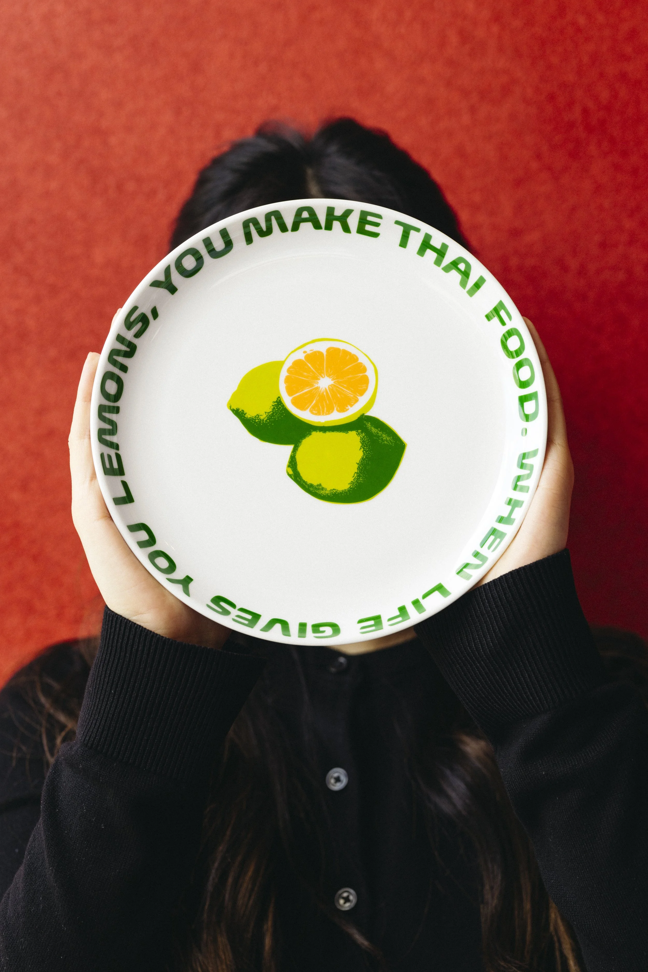 Person holding a white plate with green text around the rim saying, 'You make Thai food better,' and a graphic of two lemons, one green and one halved showing its orange interior.