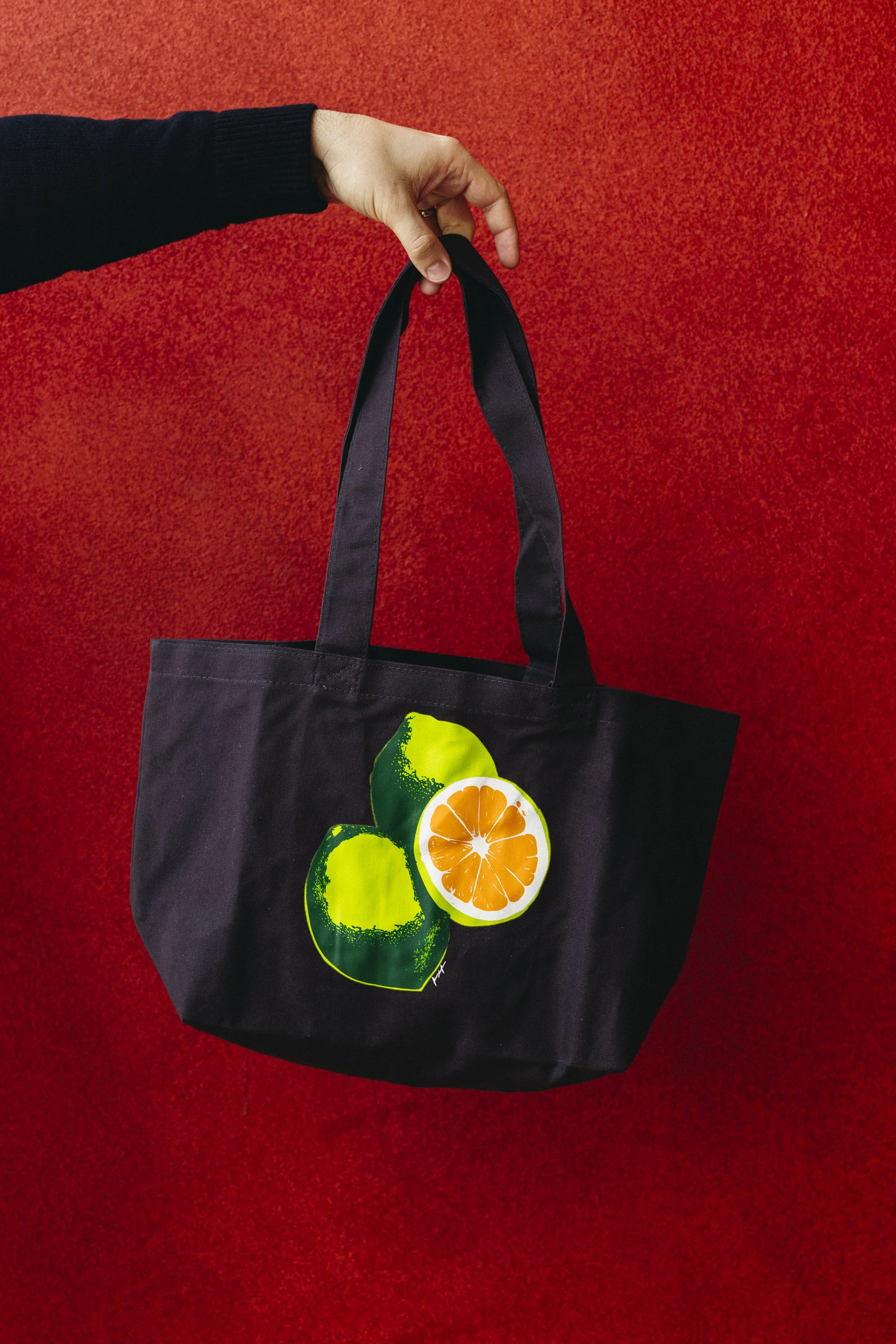 A black tote bag with citrus fruit illustrations, including lime, lemon, and orange slices, hangs from a person's hand against a red background.