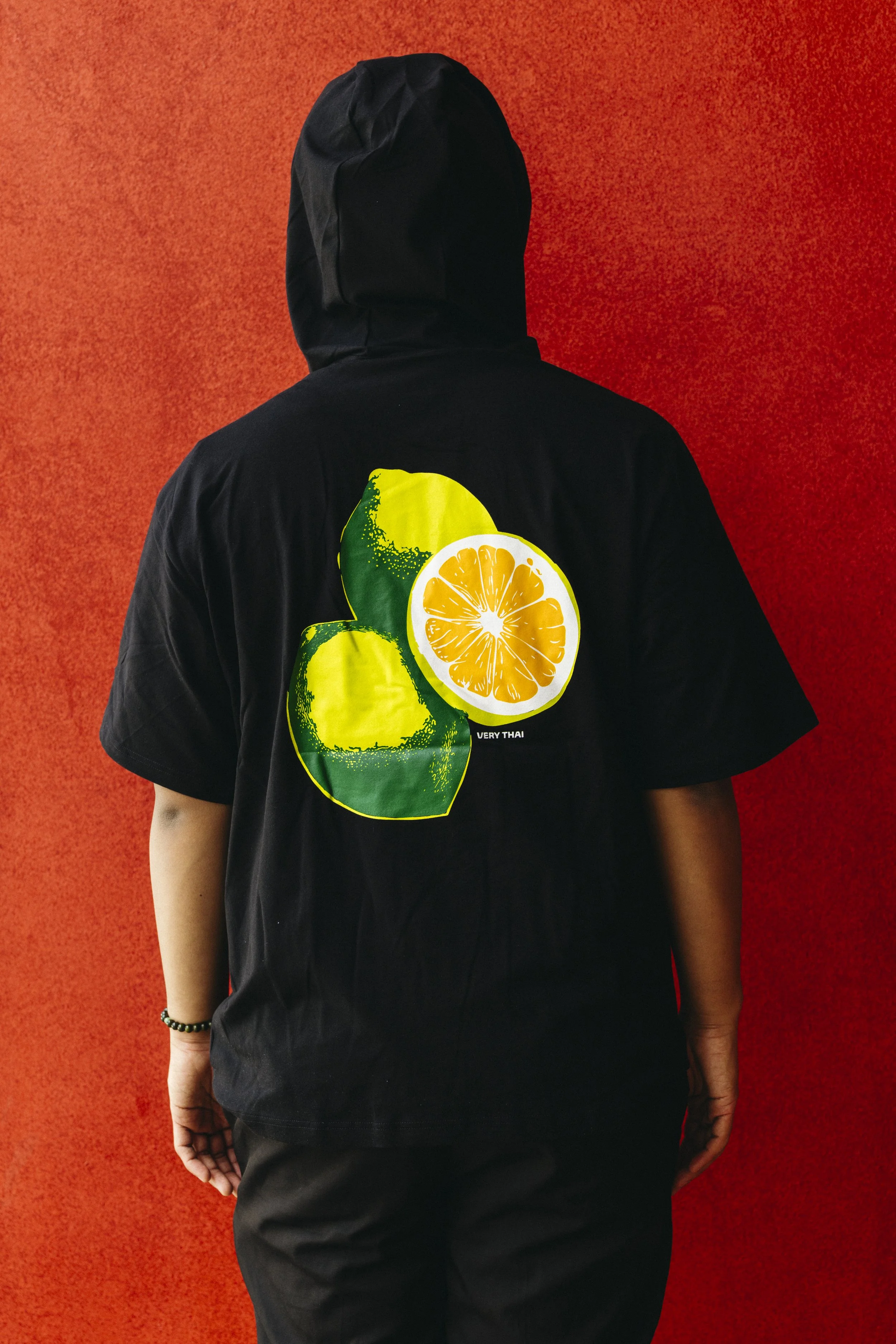 Person wearing a black t-shirt and black pants standing against a red textured background. The t-shirt has a graphic of citrus fruits, including lemons and oranges, with some text underneath.