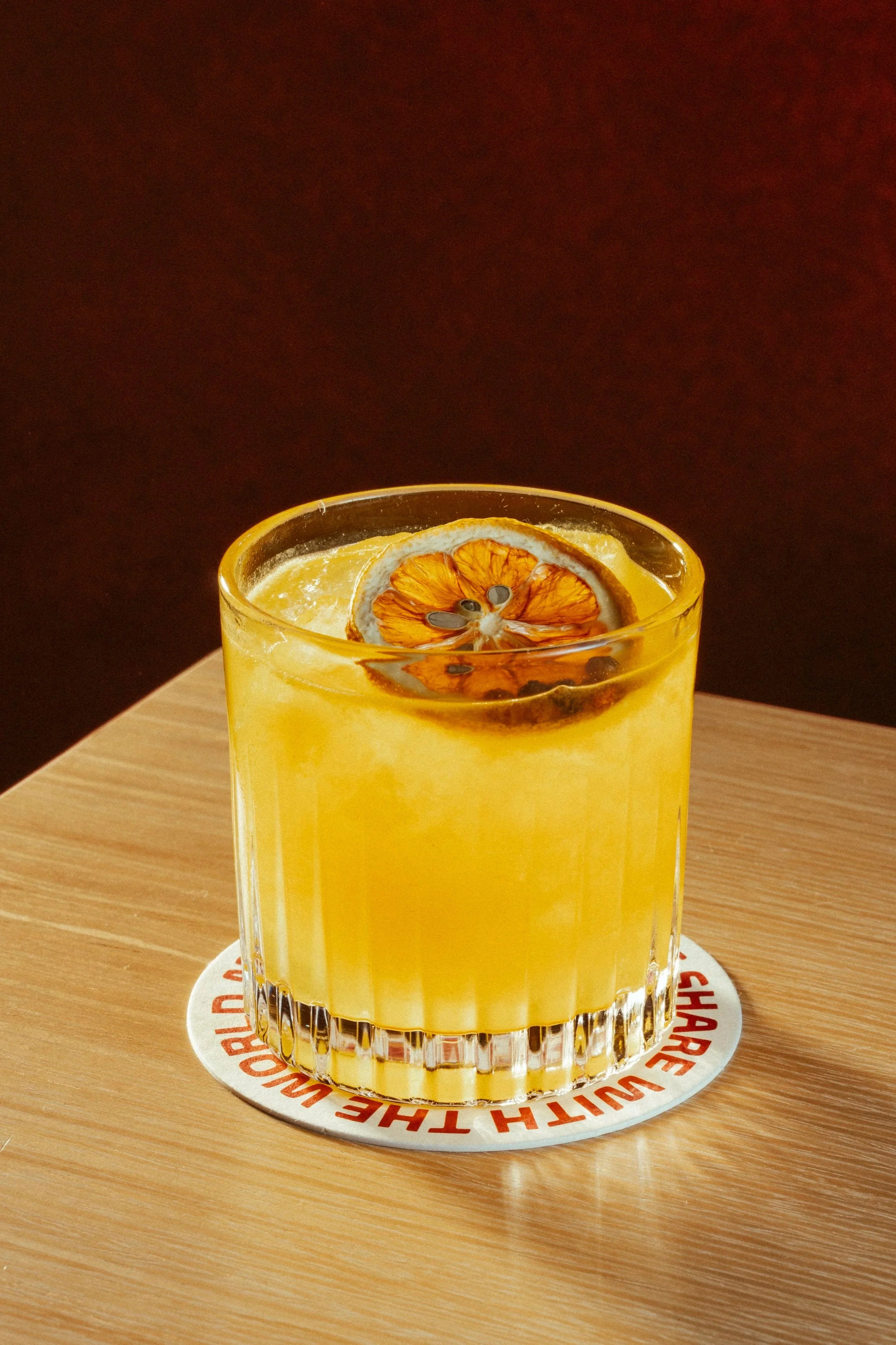 A glass of orange juice with dried orange slices on top, placed on a round charred wood coaster on a wooden table with a dark background.