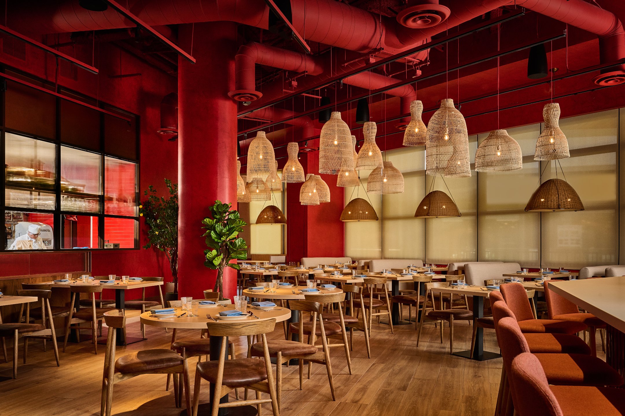 Interior of a restaurant with warm lighting, red walls, wooden furniture, hanging wicker lamps, and large windows.