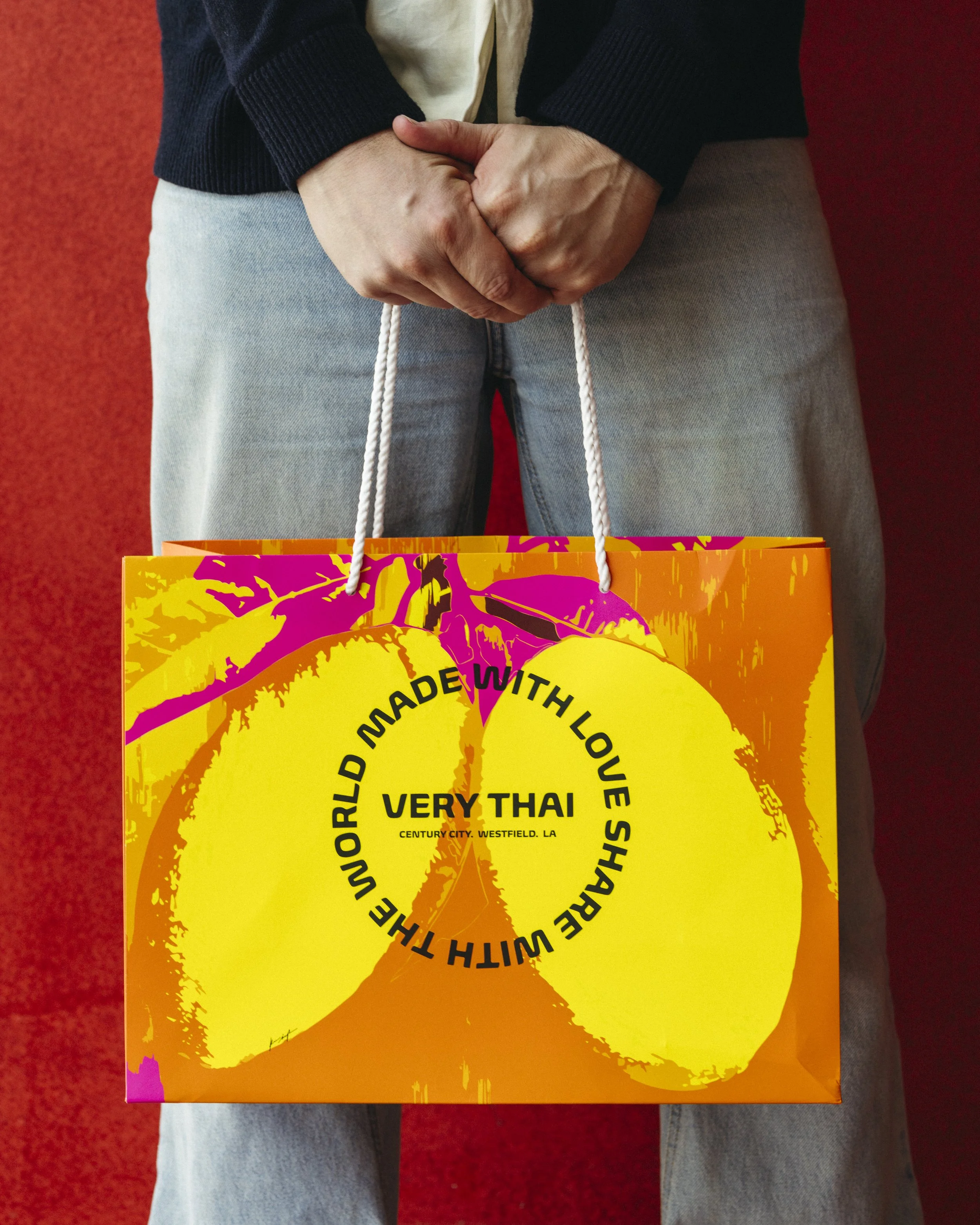 Person holding a colorful shopping bag with yellow, pink, and orange colors and text about Thai food, standing against a red background.