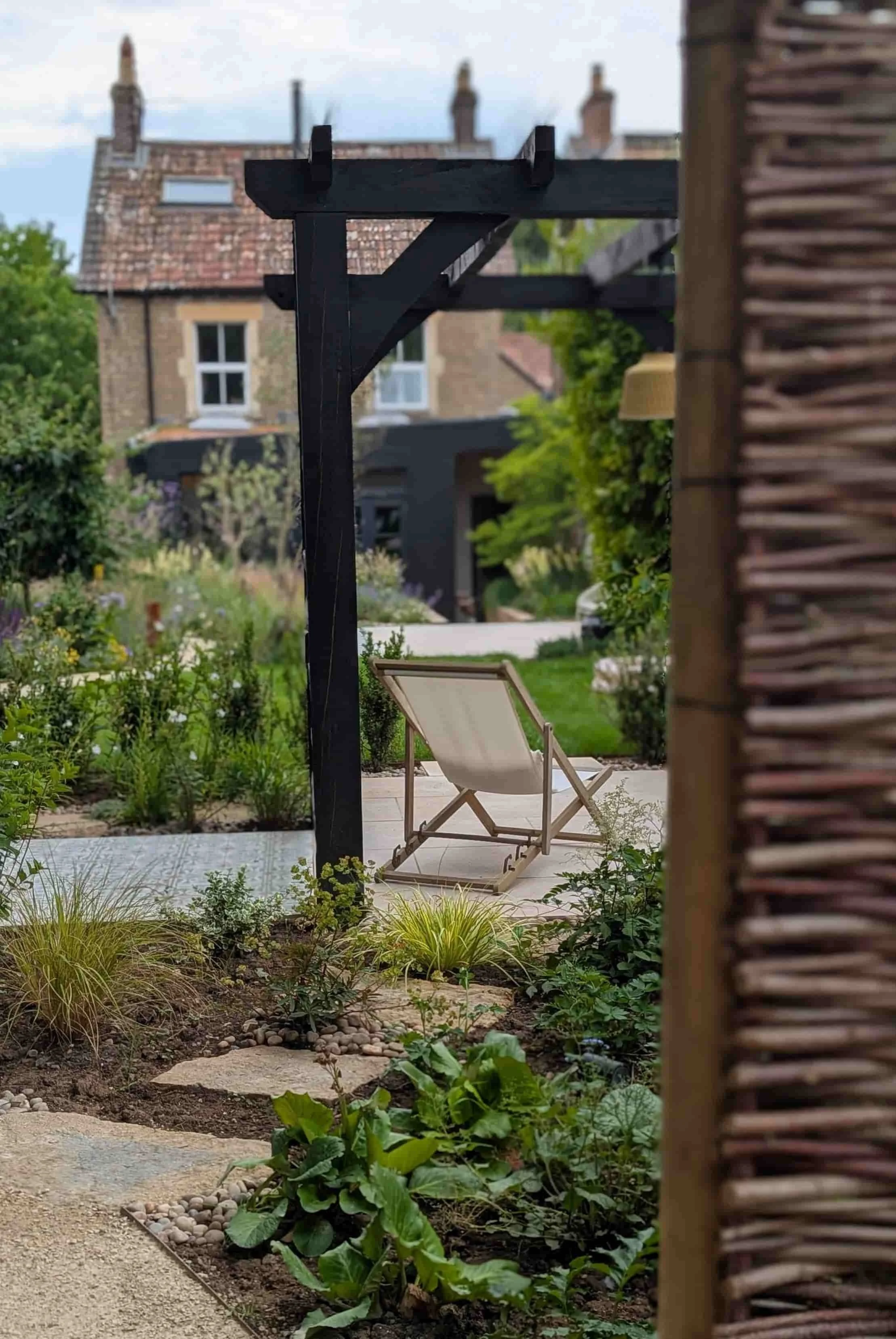 A backyard garden with a white lounge chair on a paved patio, surrounded by green plants, with a black pergola structure, and a brick house with windows in the background.