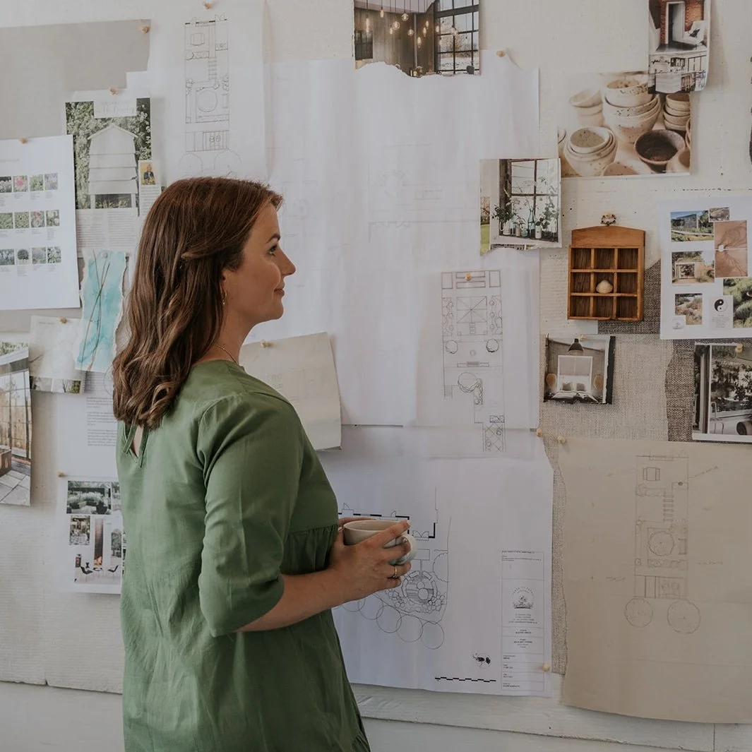 Diane Samways with brown hair wearing a green dress holding a mug, looking at a wall with architectural plans, sketches, photographs, and design ideas pinned up.