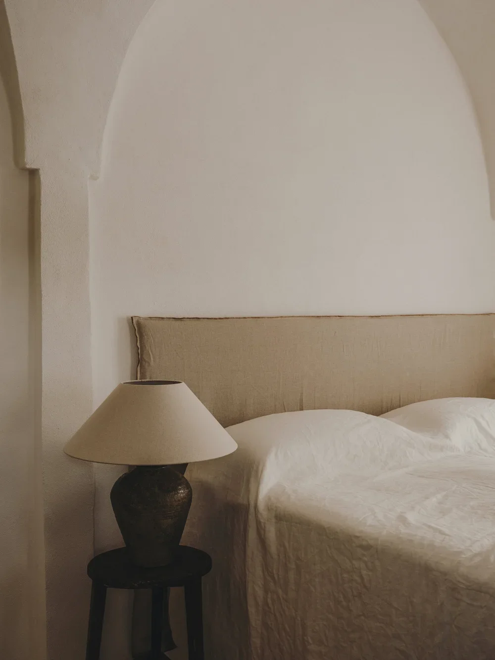 A cozy bedroom corner with a beige upholstered headboard, white bedding, a wooden side table, and a table lamp in a dark textured finish with a beige lampshade.