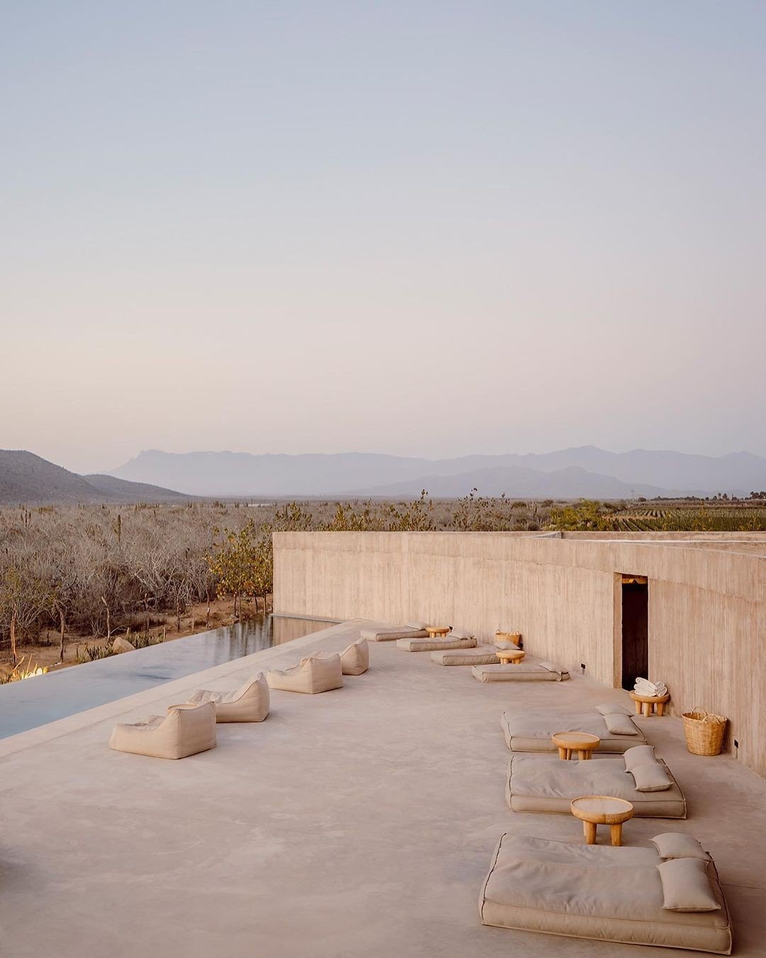 An outdoor terrace with modern beige seating cushions and small round wooden tables, overlooking a desert landscape with mountains in the distance.