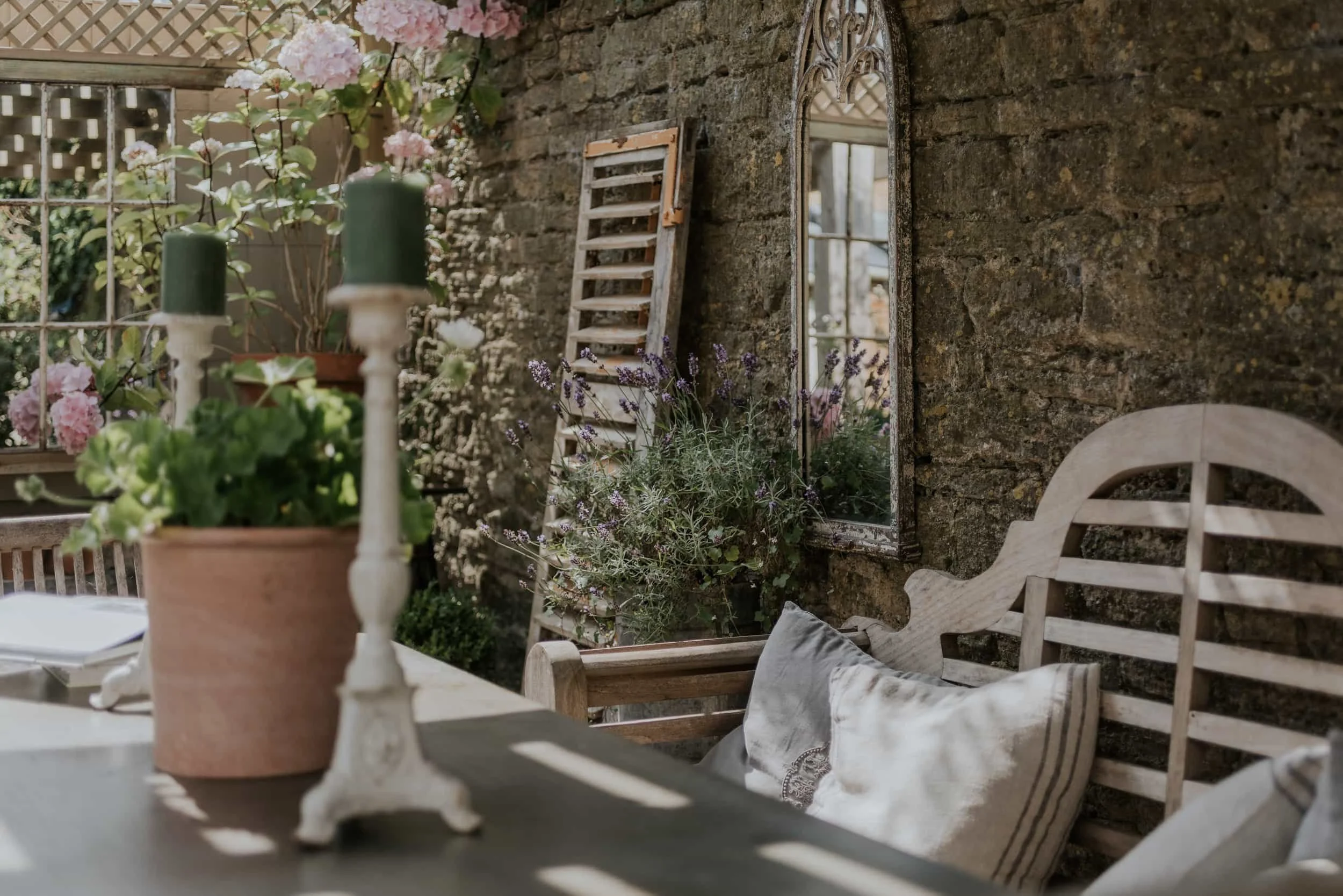A rustic outdoor patio with a stone wall, a mirror, potted plants, and cushions on a wooden bench, illuminated by natural light.