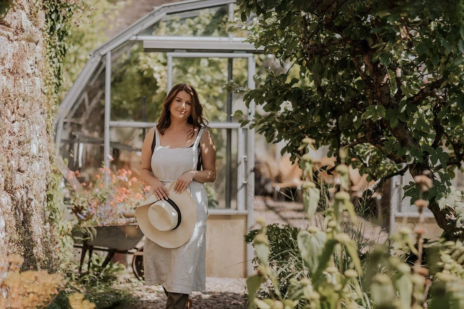 Diane Samways walking in a garden holding a straw hat, with greenery and a greenhouse in the background.