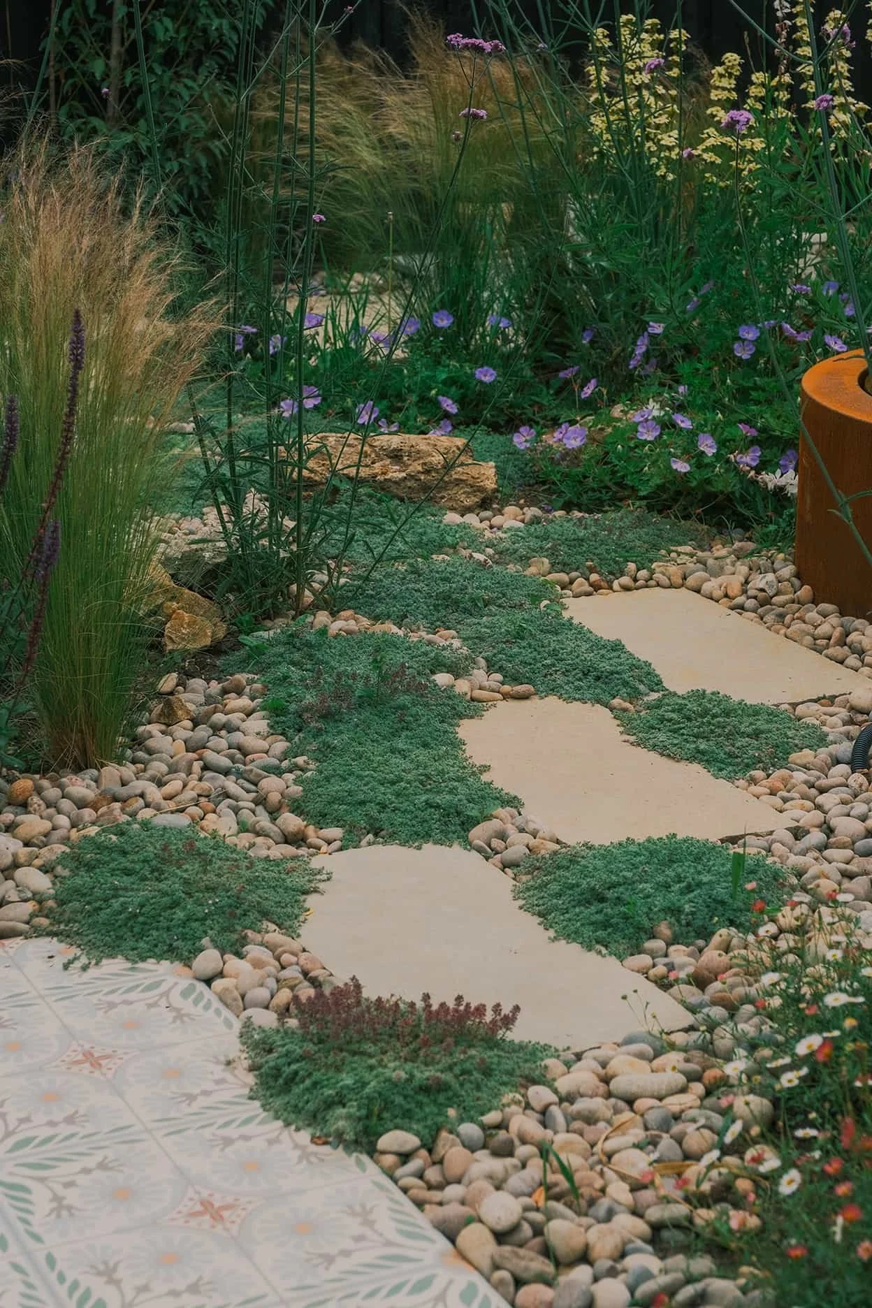 A garden path with irregular stepping stones surrounded by green ground cover, decorative rocks, and flowering plants including purple and white flowers.