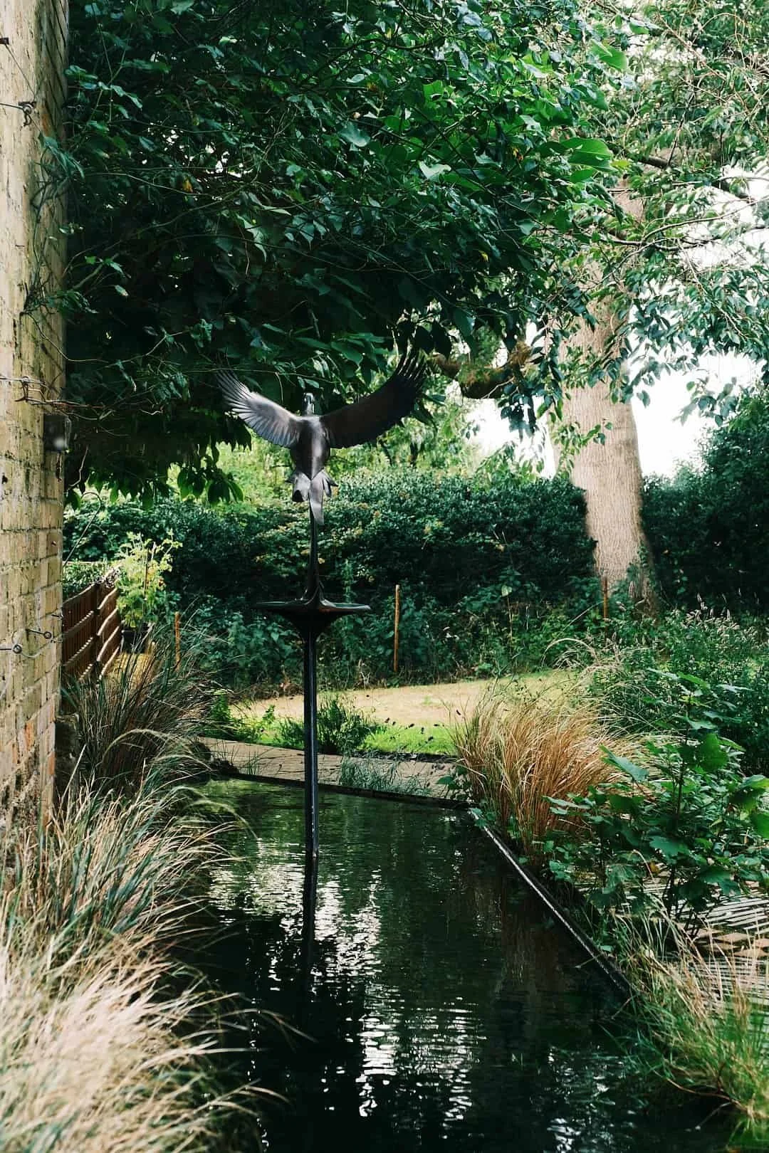 A garden with a narrow water feature, ornamental grasses, lush green bushes, and trees. A bird sculpture with wings spread is mounted on a pole above the water.