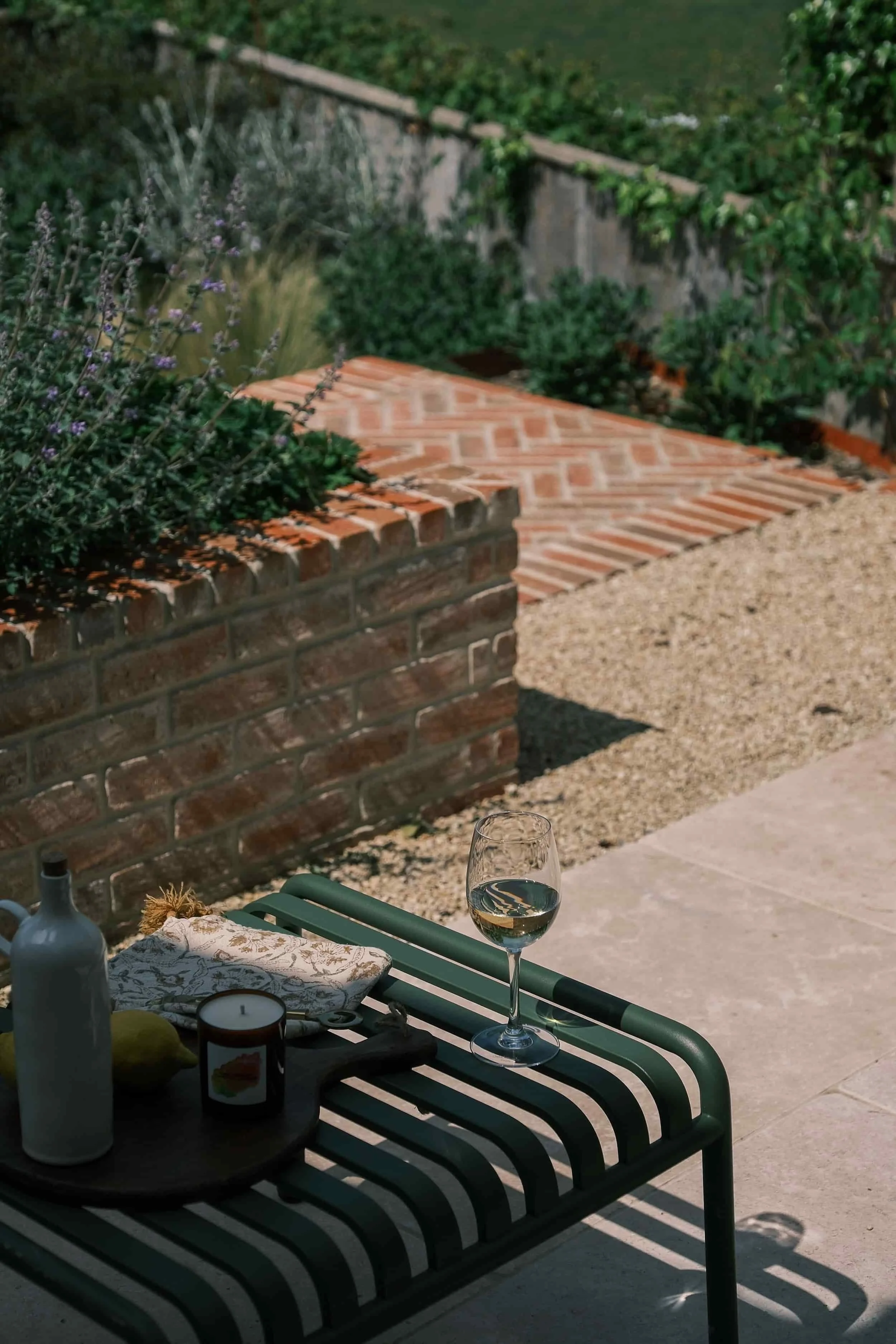 Outdoor patio scene featuring a glass of white wine on a black and white striped bench, with a bottle, candle, and a cloth napkin, against a backdrop of a brick wall, gravel, and lush greenery.