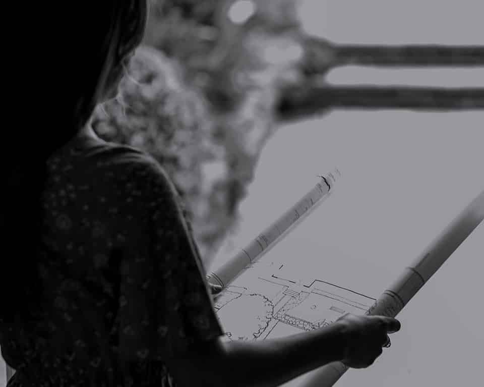 A young girl with curly hair wearing a patterned shirt drawing architectural plans for a Garden Design on a large sheet of paper.