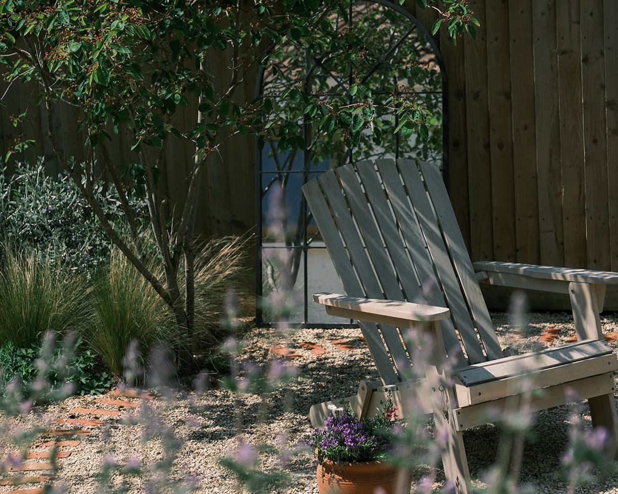 A garden scene with two wooden Adirondack chairs, some purple flowers in a pot, and greenery including bushes and trees, enclosed by a wooden fence.