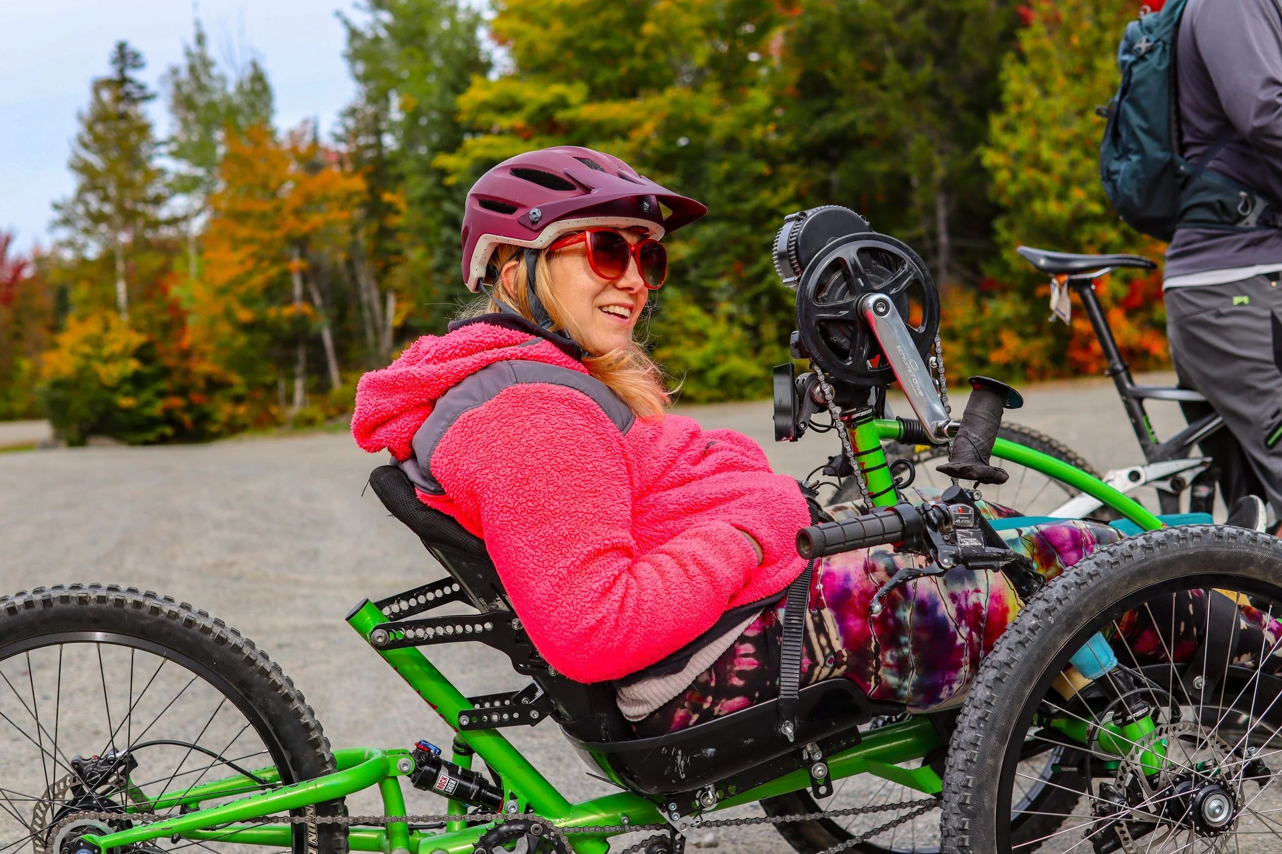 Smiling woman in a helmet and red jacket riding a green adaptive handcycle outdoors, surrounded by trees with fall foliage.