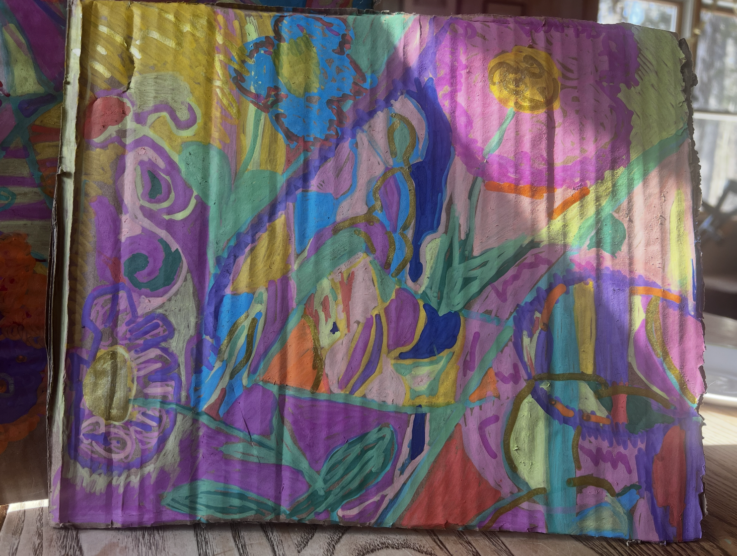 Colorful abstract painting on paper with various shapes and brushstrokes in purple, blue, green, pink, yellow, orange, and gold, displayed on a wooden table.