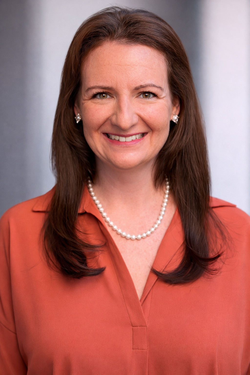 A woman with brown hair wearing a rust-colored blouse, pearl necklace, and pearl earrings, smiling.