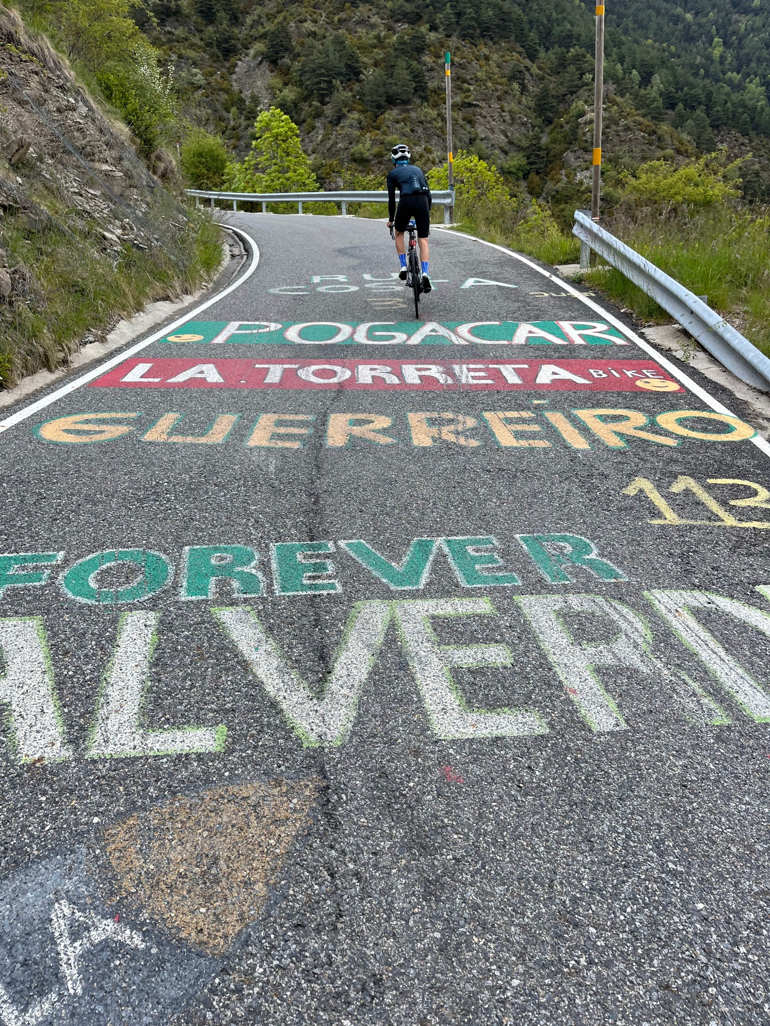 A cyclist riding up a mountain road decorated with colorful chalk art and text that says 'Guerra', 'Forever', and other phrases, with lush green trees and rocky hills in the background.