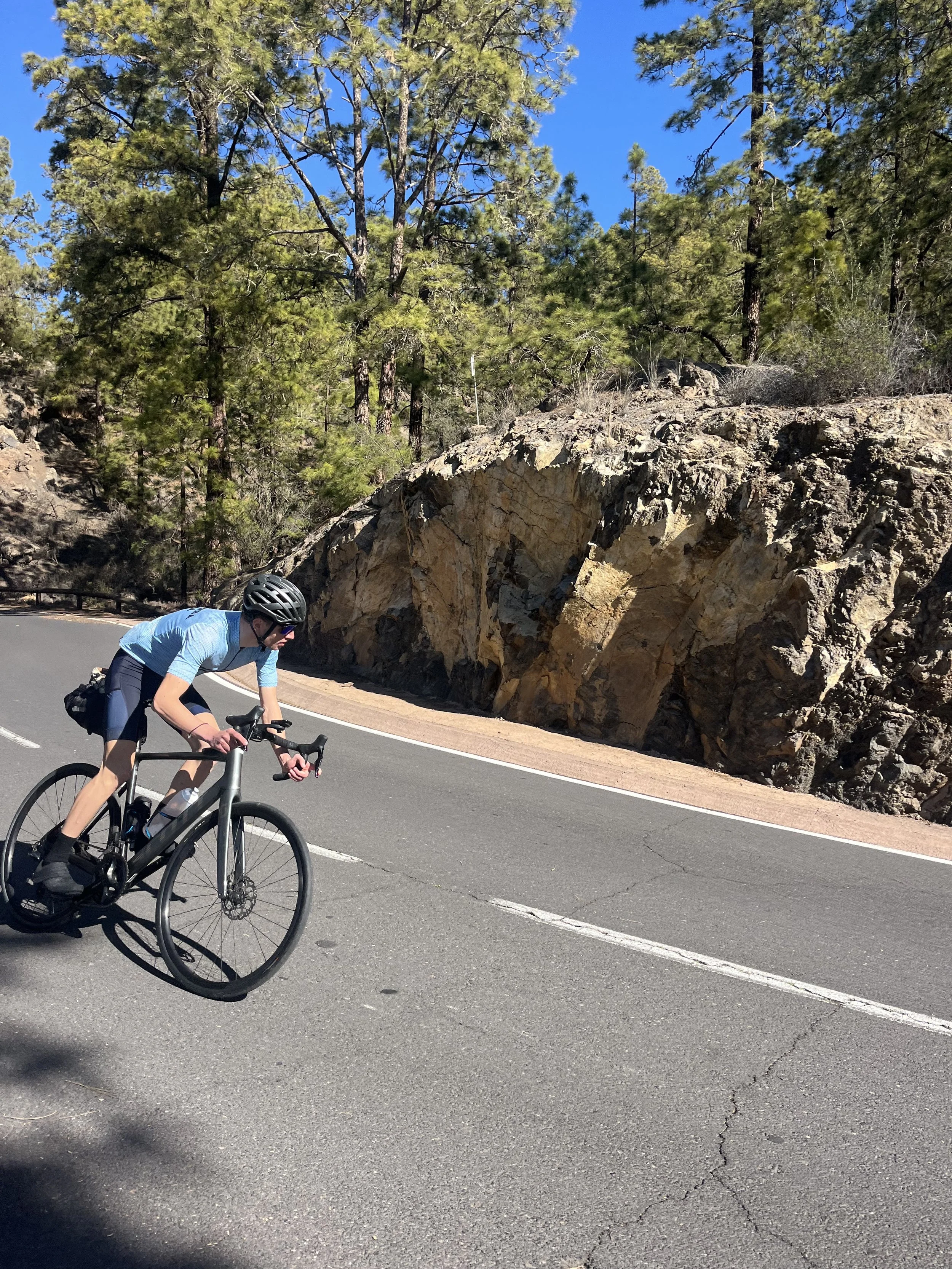 A person riding a bicycle on a mountain road surrounded by trees and large rocks under a clear blue sky.