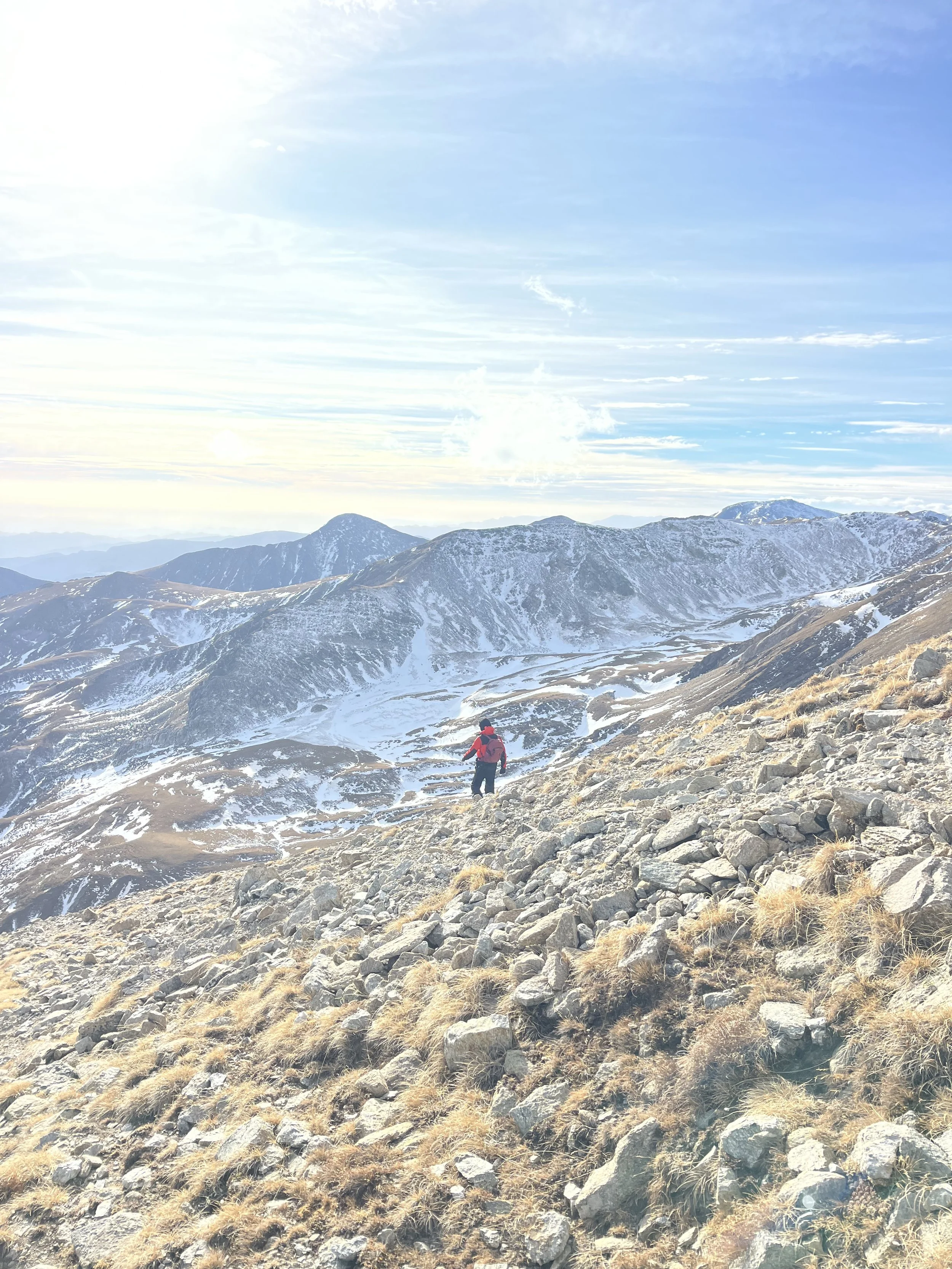 A person hiking on a rocky mountain with snow-capped peaks in the background under a partly cloudy sky.