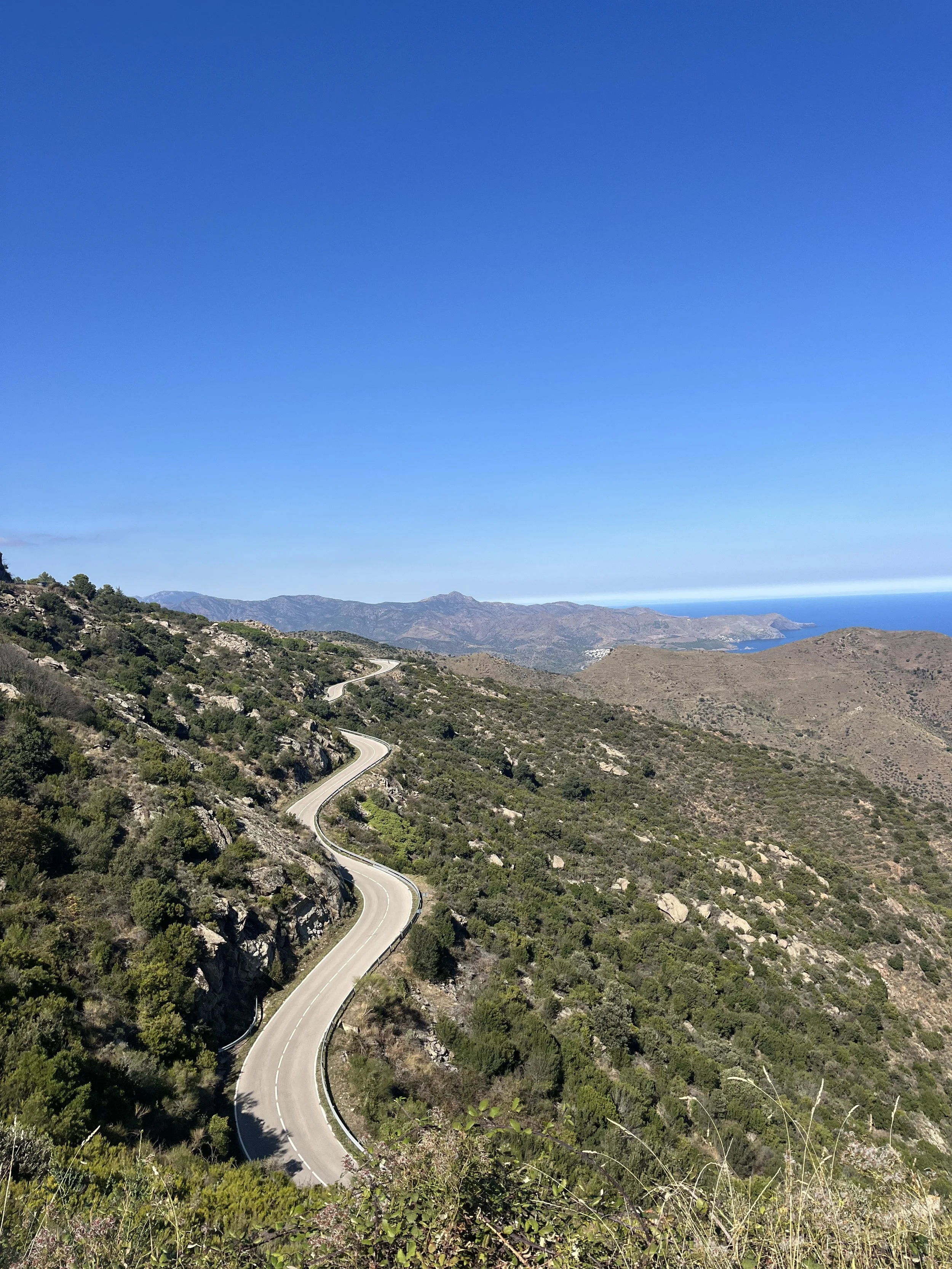 A winding mountain road with guardrails, surrounded by green shrubbery and rocky terrain, under a clear blue sky with distant mountains and the ocean on the horizon.