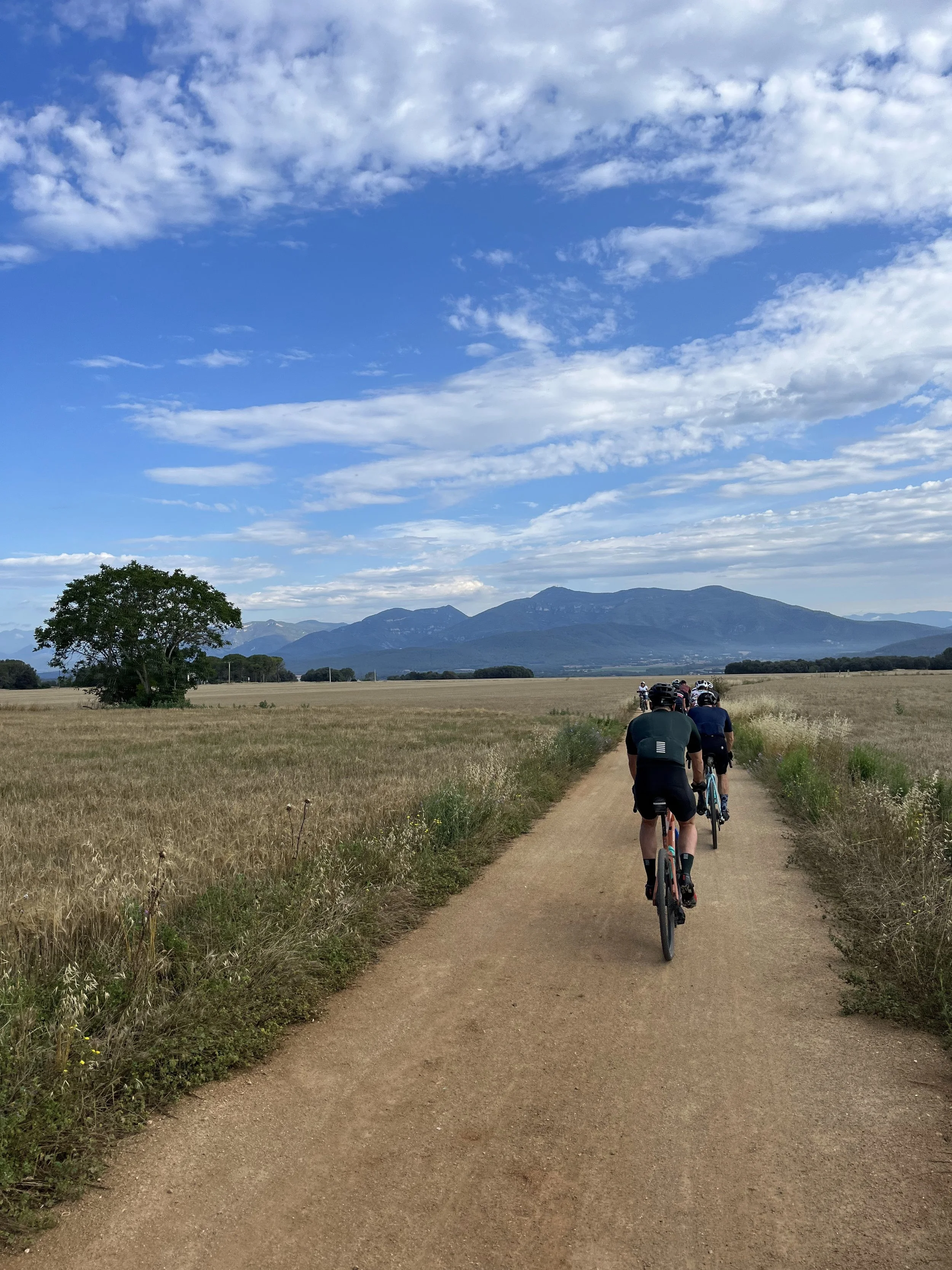 A group of cyclists riding on a dirt path through an open field with mountains in the background under a partly cloudy sky.