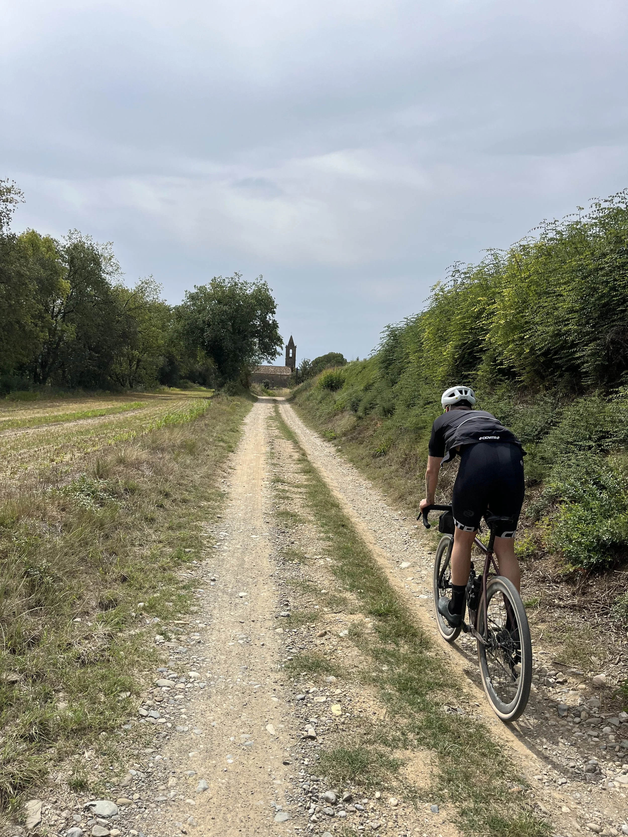 A person riding a gravel bike on a dirt trail in a rural area with fields and trees on either side, and a church steeple in the distance under a cloudy sky.