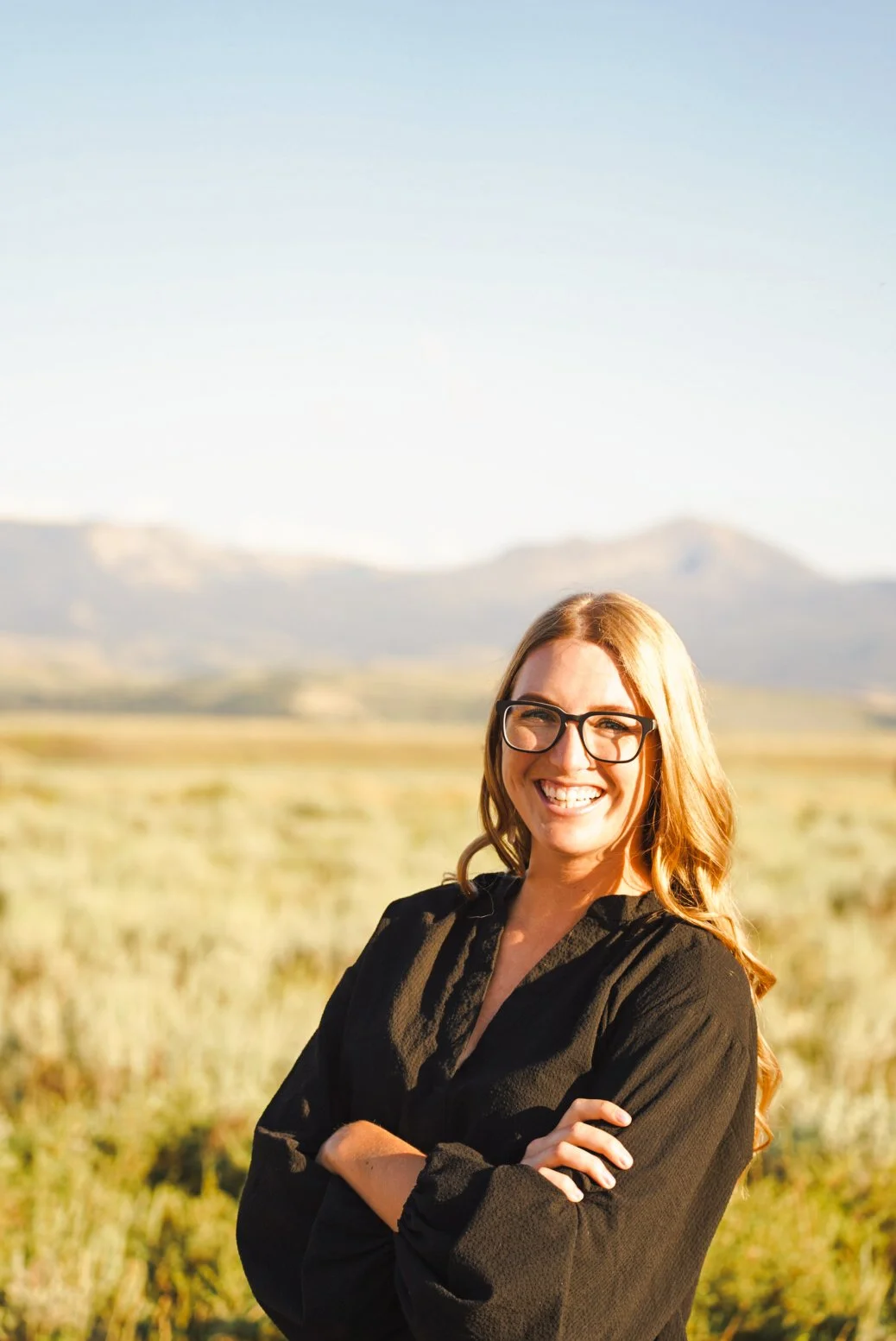 A woman with blonde hair, glasses, and a black shirt smiling with arms crossed outdoors, with mountains in the background under a clear sky.