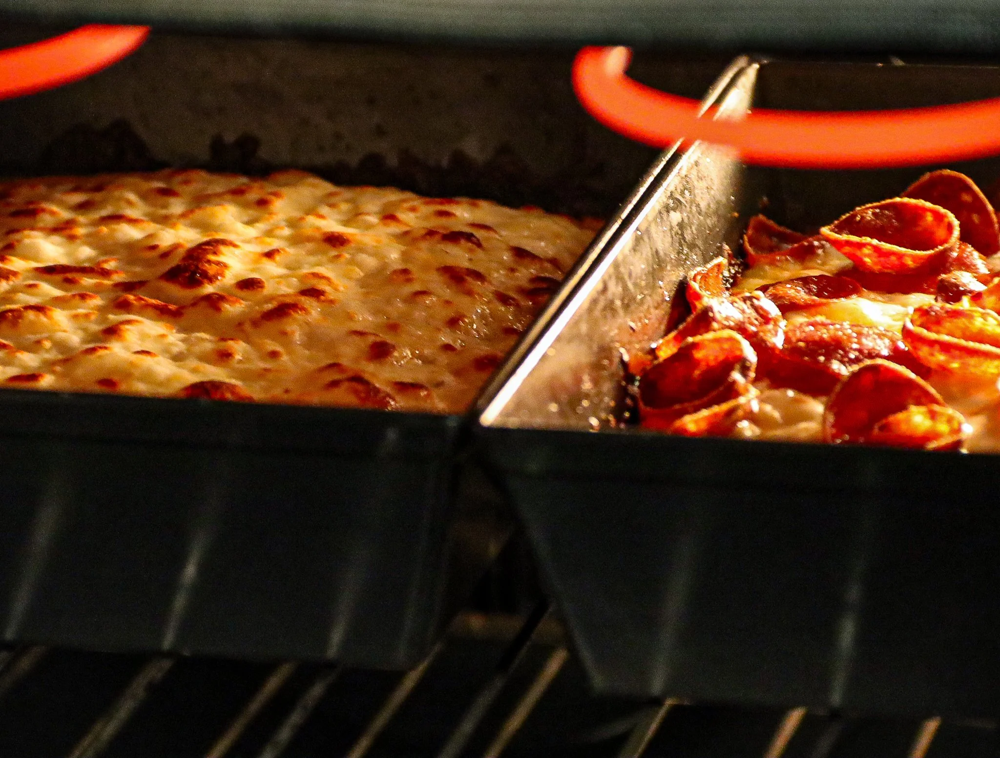 Close-up of two Detroit-style pizzas baking in an oven in rectangular pans, one cheese and the other with pepperoni.