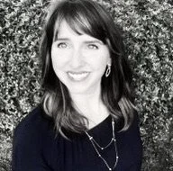 Smiling woman with shoulder-length dark hair, earrings, and a dark top standing outdoors in front of foliage, black-and-white photo.