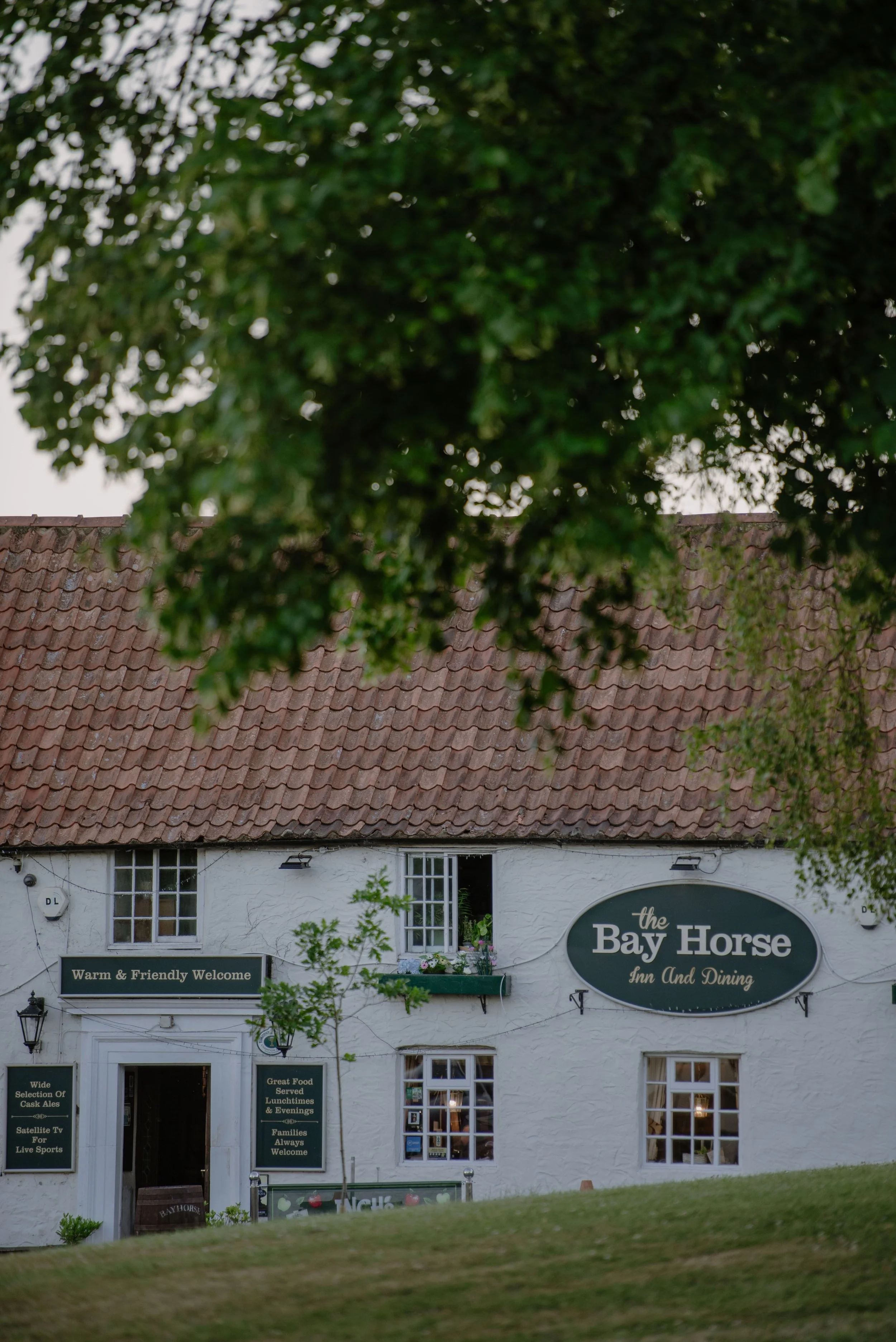 A white building with a red tiled roof, green signs, and an oval sign that reads 'the Bay Horse Inn And Dining.' There are small windows, some with flower boxes, and a grassy foreground surrounds the building.