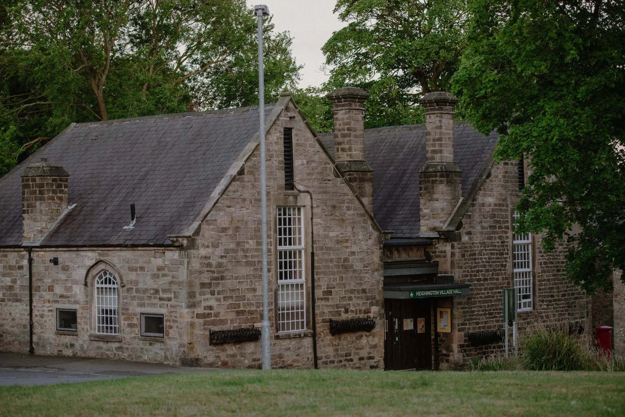 A stone building with arched windows and a slate roof, surrounded by trees, with a street sign that reads 'Heinghamton Village Hall.'