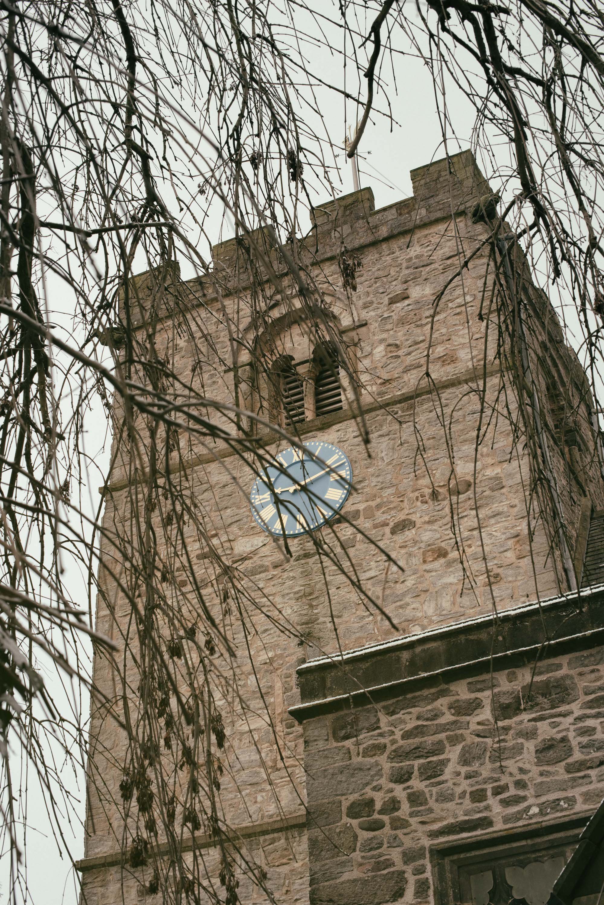 A stone clock tower with a round clock face showing the time 12:50, surrounded by barren tree branches, on a cloudy day.