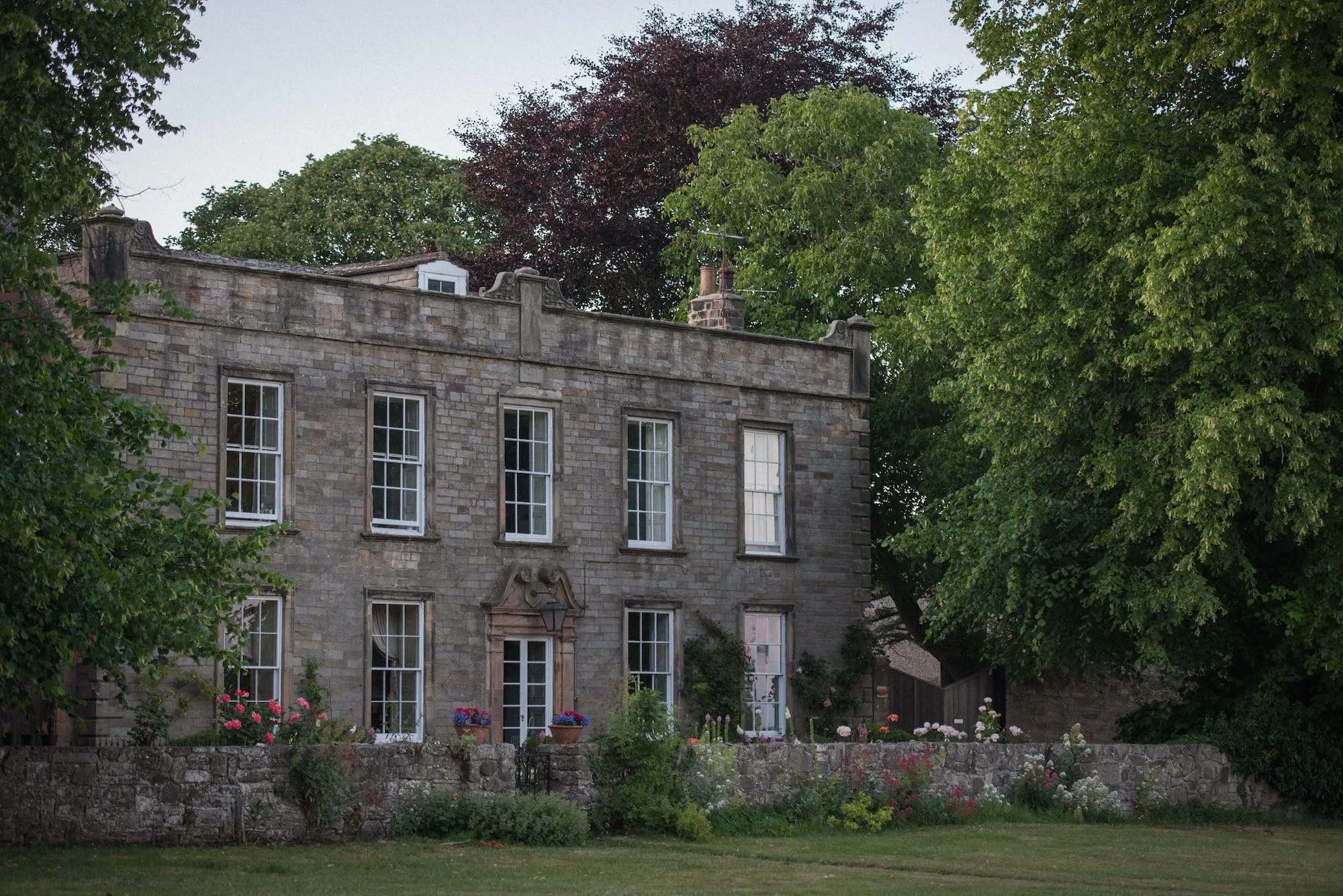A historic stone building with large windows, surrounded by a garden with flower pots and a lawn, flanked by tall green trees.