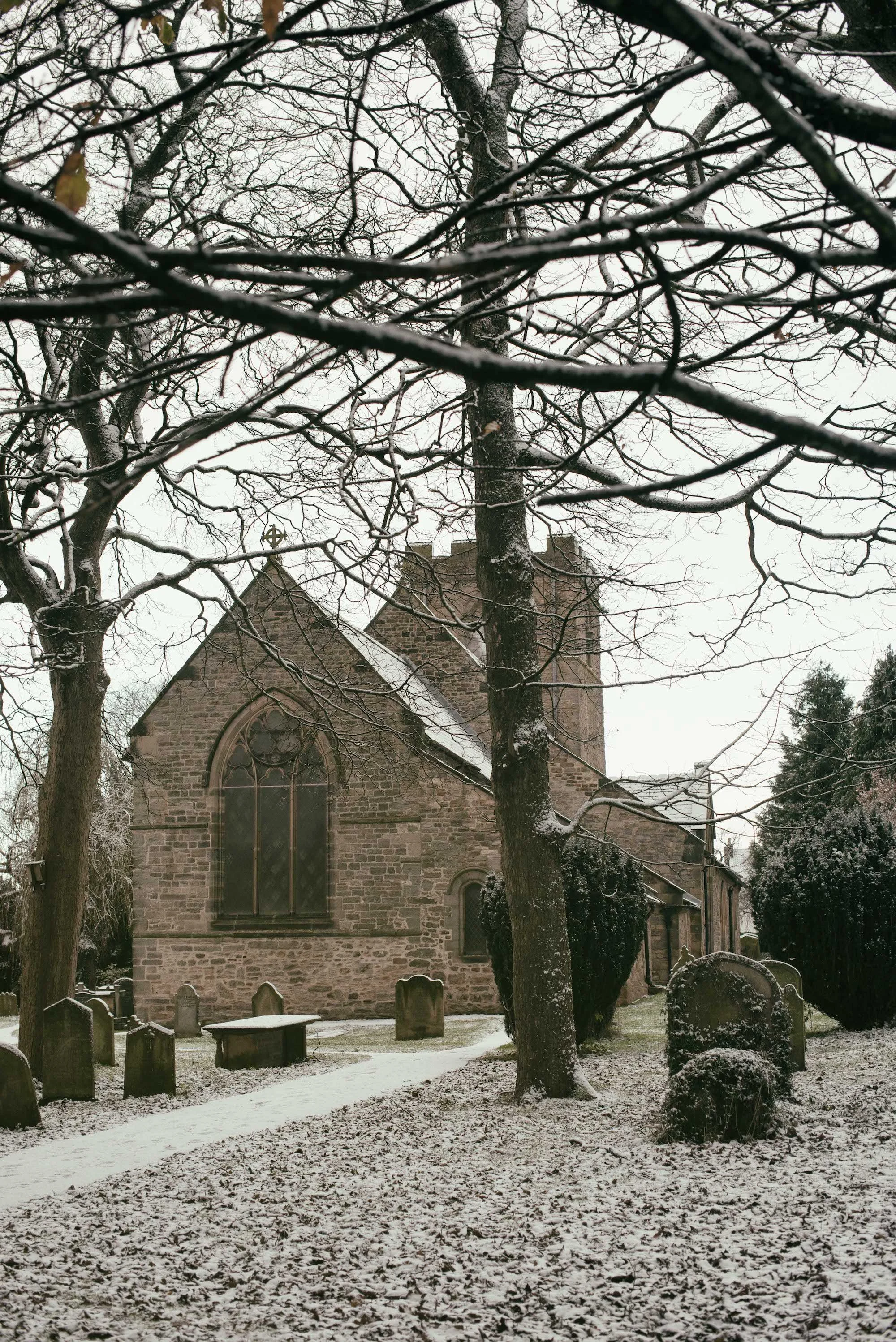 A stone church with arched windows seen behind snow-covered trees and a graveyard with tombstones, in a winter setting.