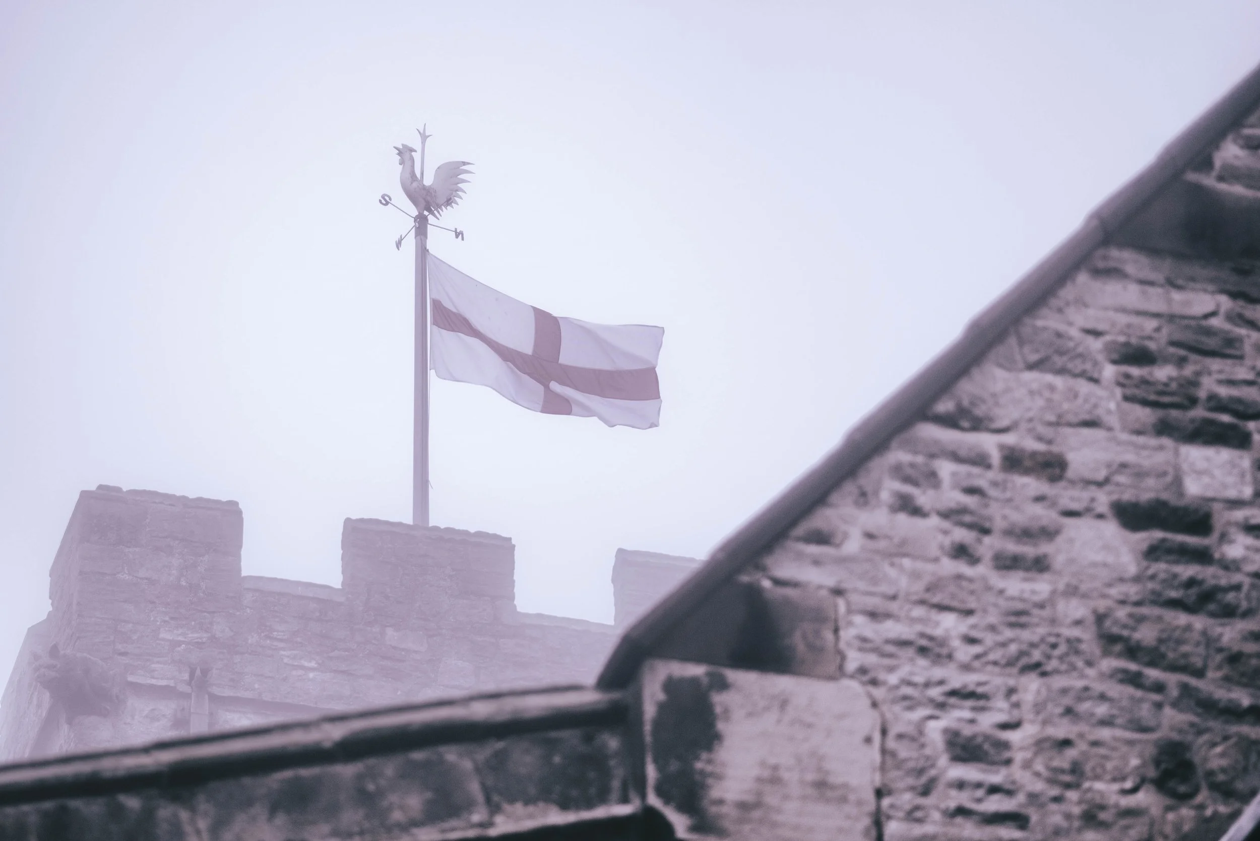A weathervane with a rooster figure on top of a stone castle wall and an Irish flag flying nearby, under a cloudy sky.