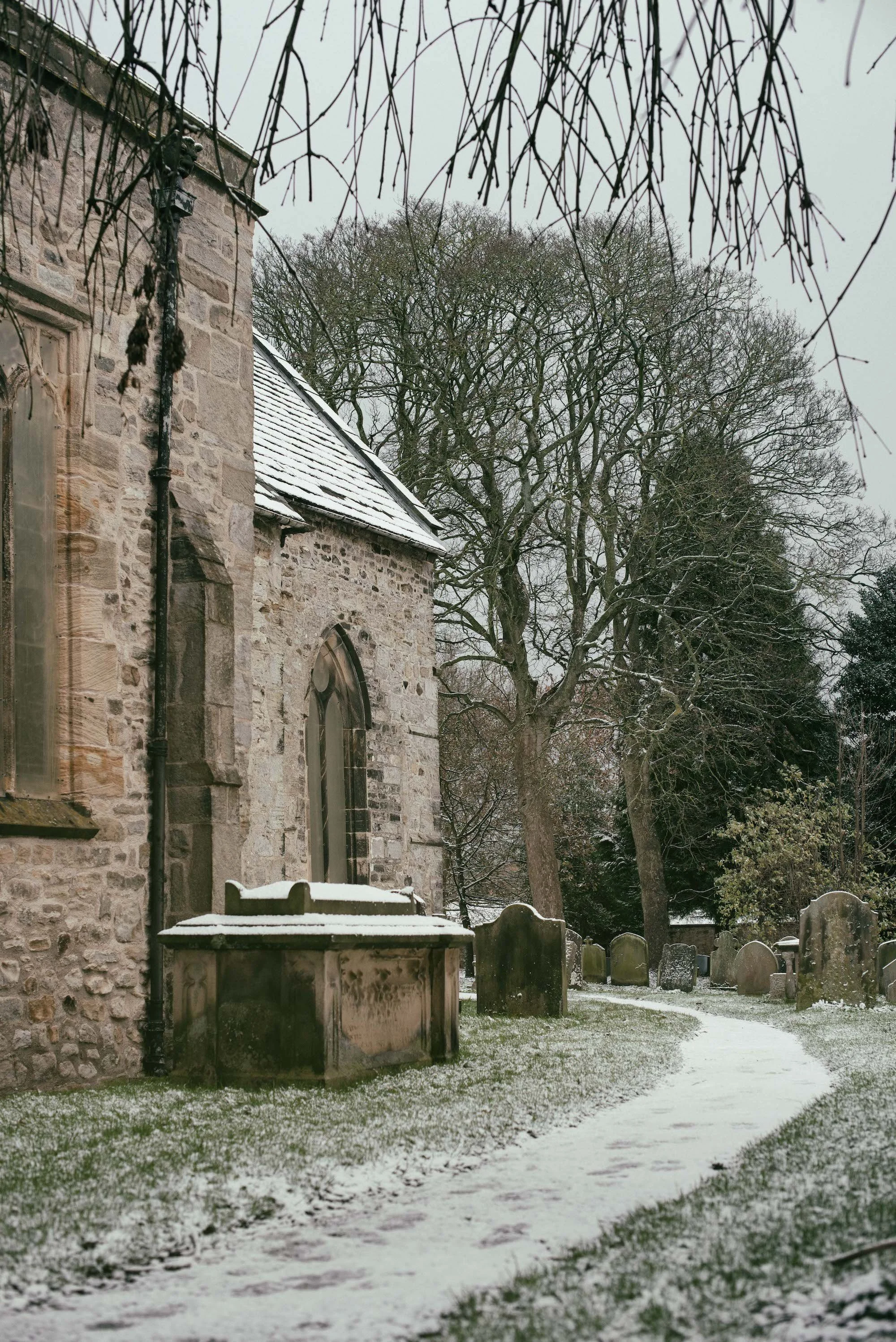 A stone church with arched windows and a metal roof, surrounded by leafless trees and old gravestones in a snow-covered graveyard.