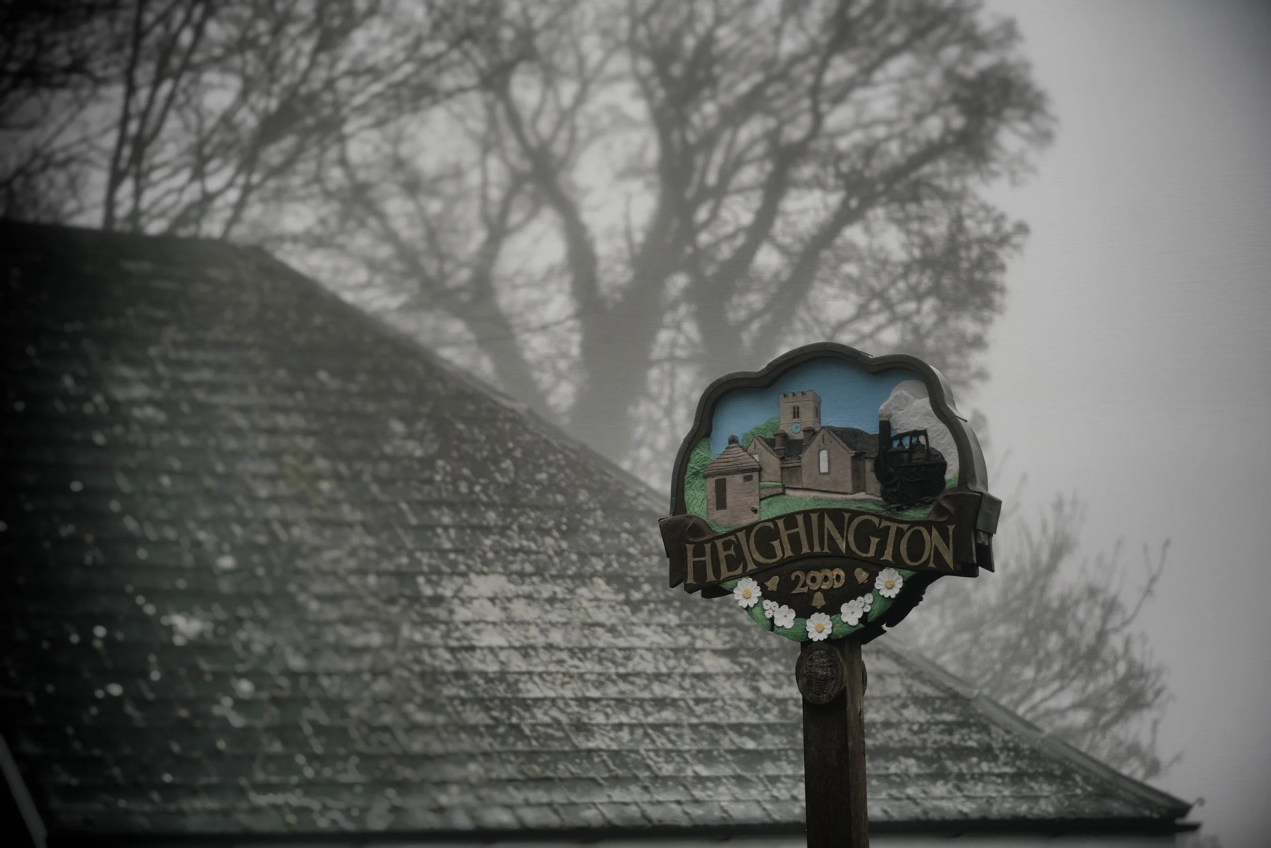 A decorative sign for Heighington village, with a painted image of a church and landscape, set against a cloudy, tree-filled background.