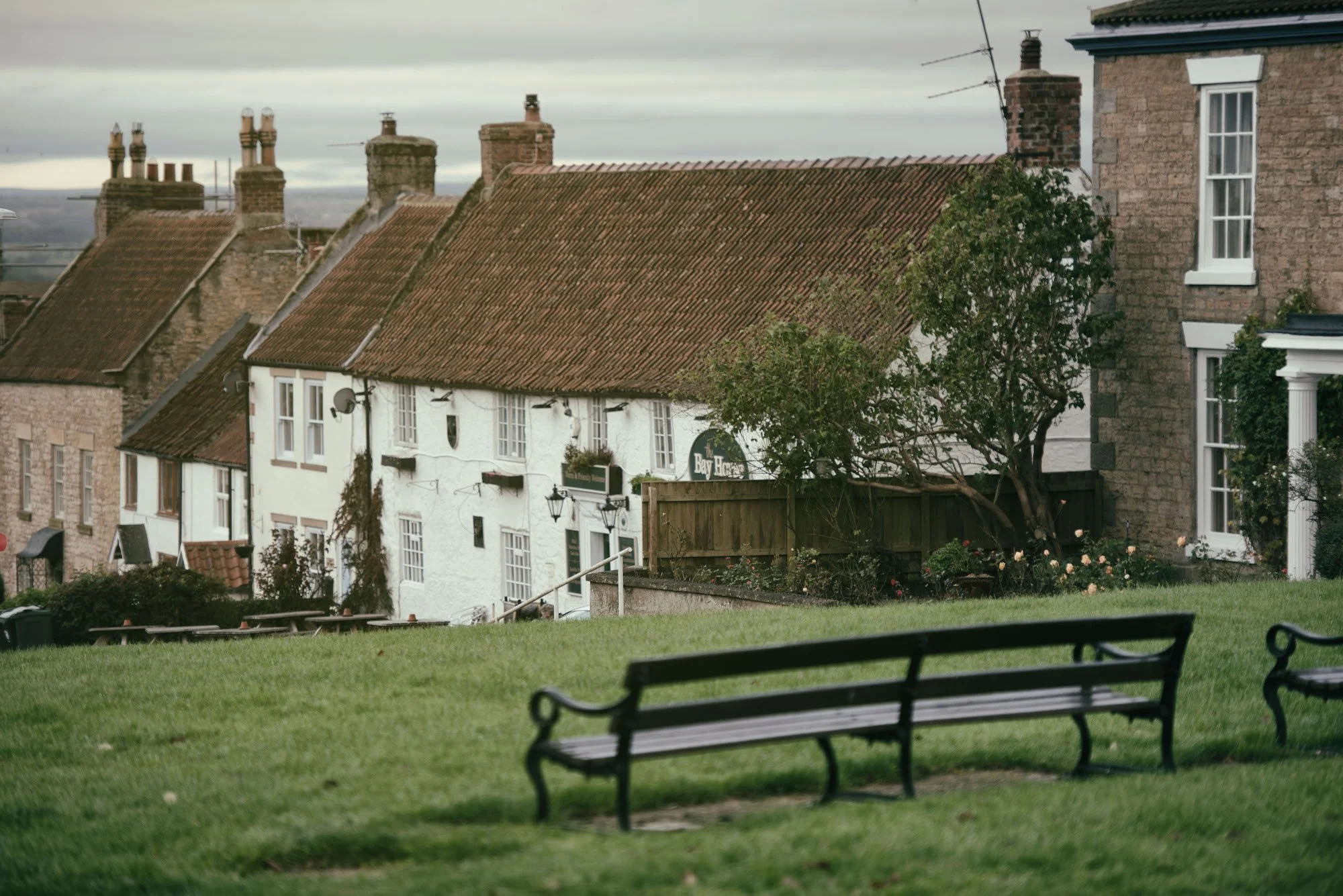 A quiet neighborhood in Heighington with stone and brick houses featuring red tiled roofs, seen from the Green with benches and a grassy area, under a cloudy sky.